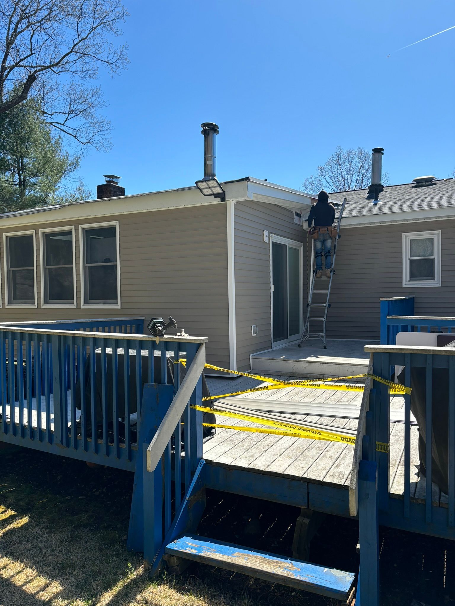 A person on a ladder near a house roof. A deck with blue railings is in the foreground. Sunny, daytime.