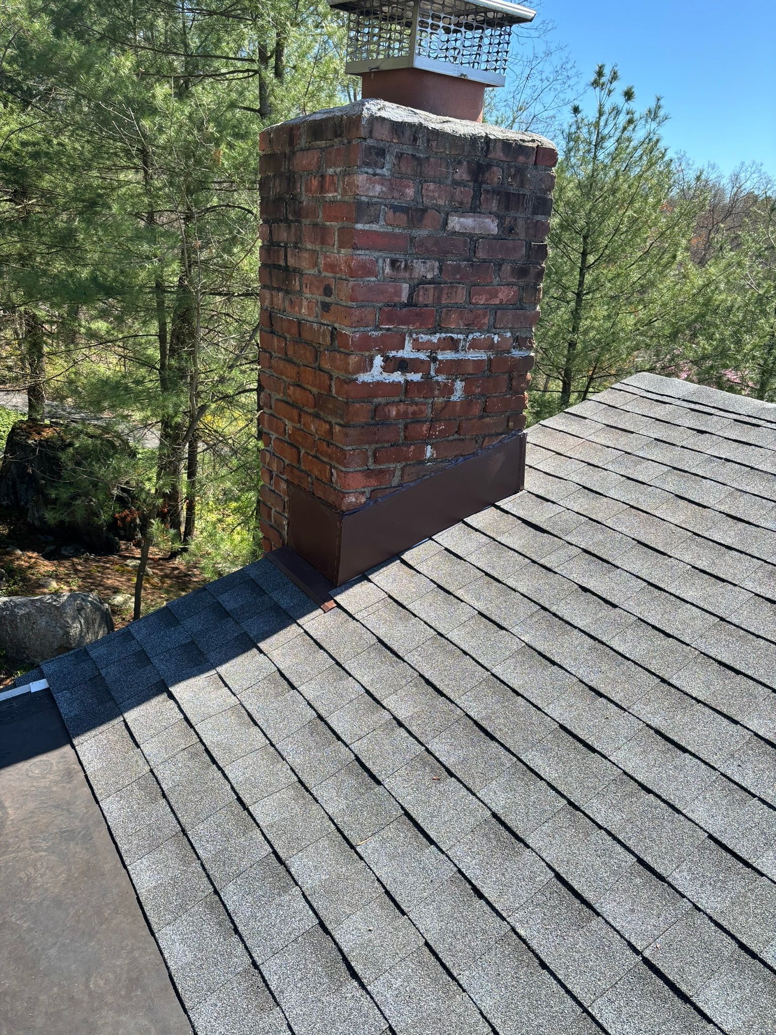 Brick chimney on a shingled roof, weathered bricks, surrounded by trees, clear sky.