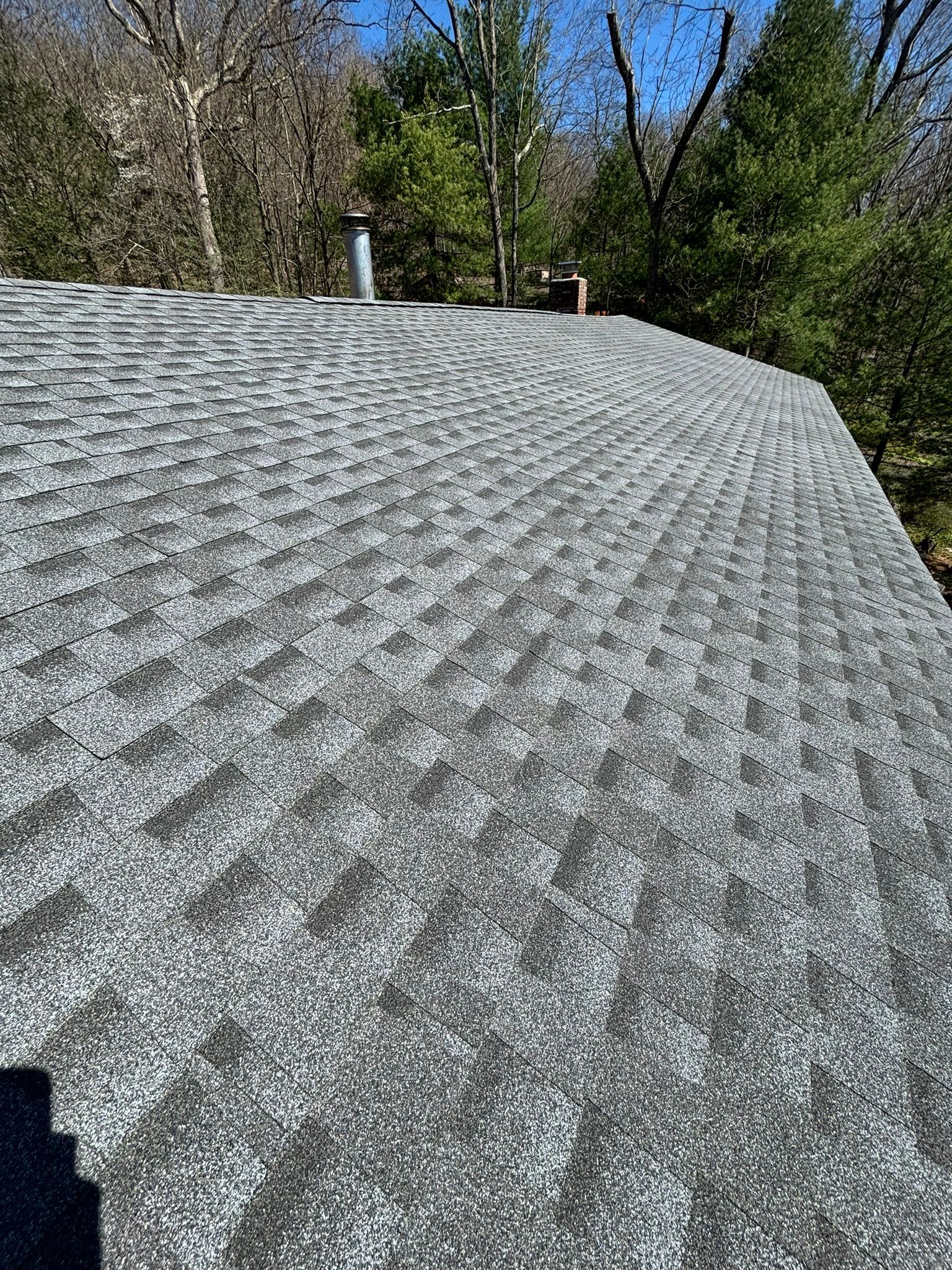 Gray asphalt shingle roof with metal chimney against a background of trees and blue sky.