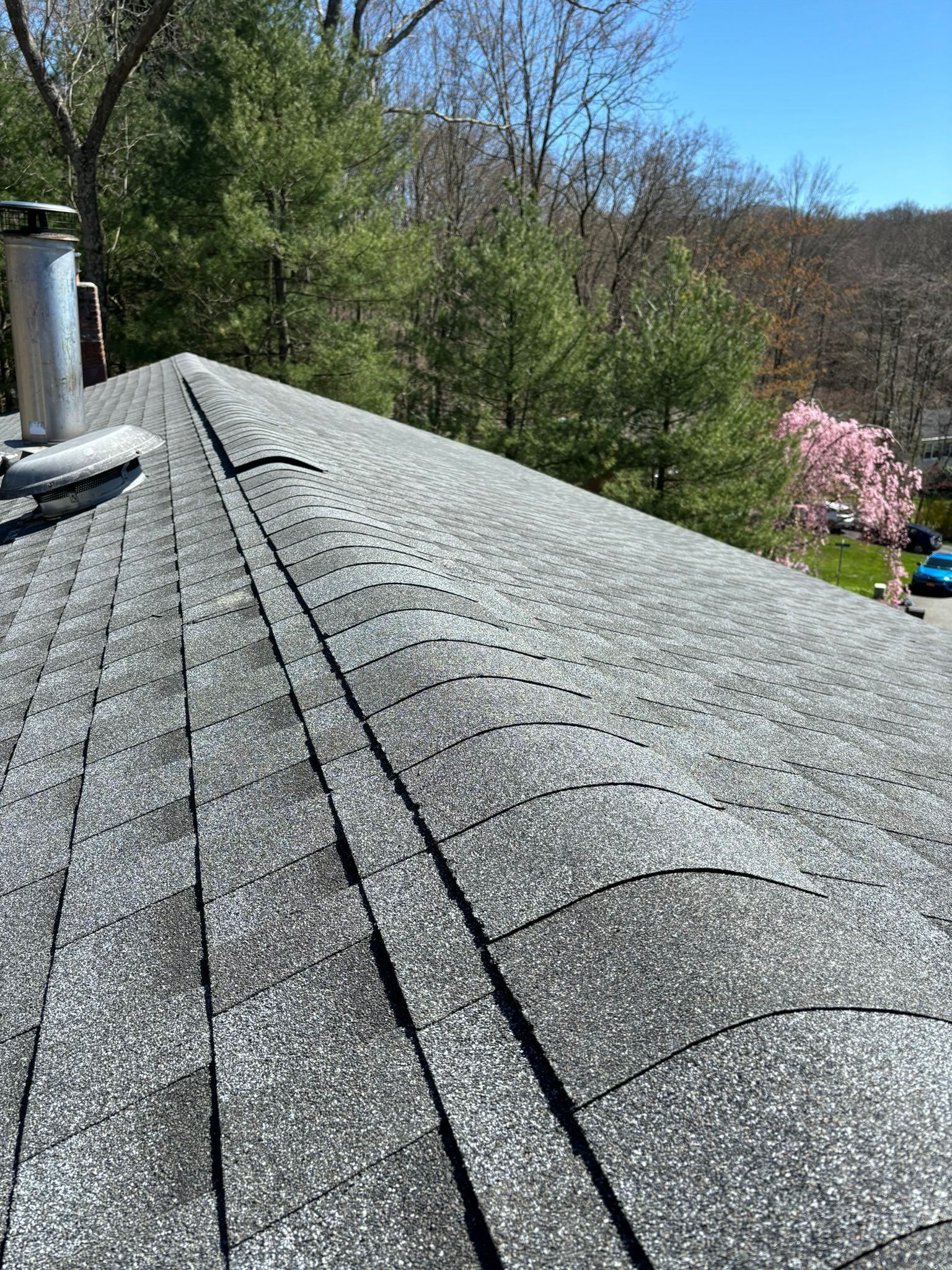 Gray asphalt shingle roof with a chimney against a backdrop of trees and a blue sky.