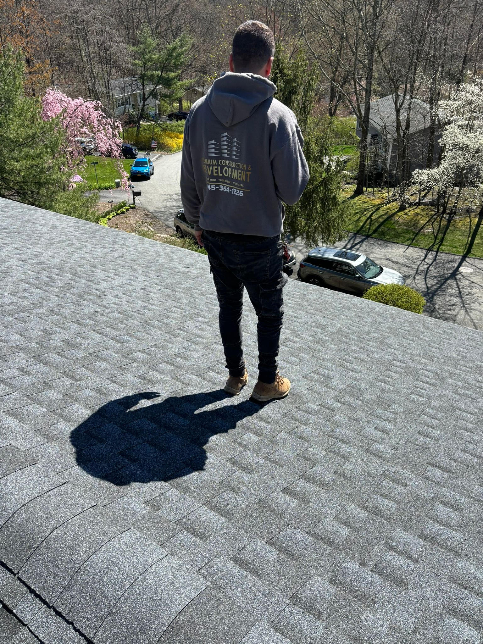 Man standing on a gray shingle roof, casting a shadow. Houses and trees in the background on a sunny day.