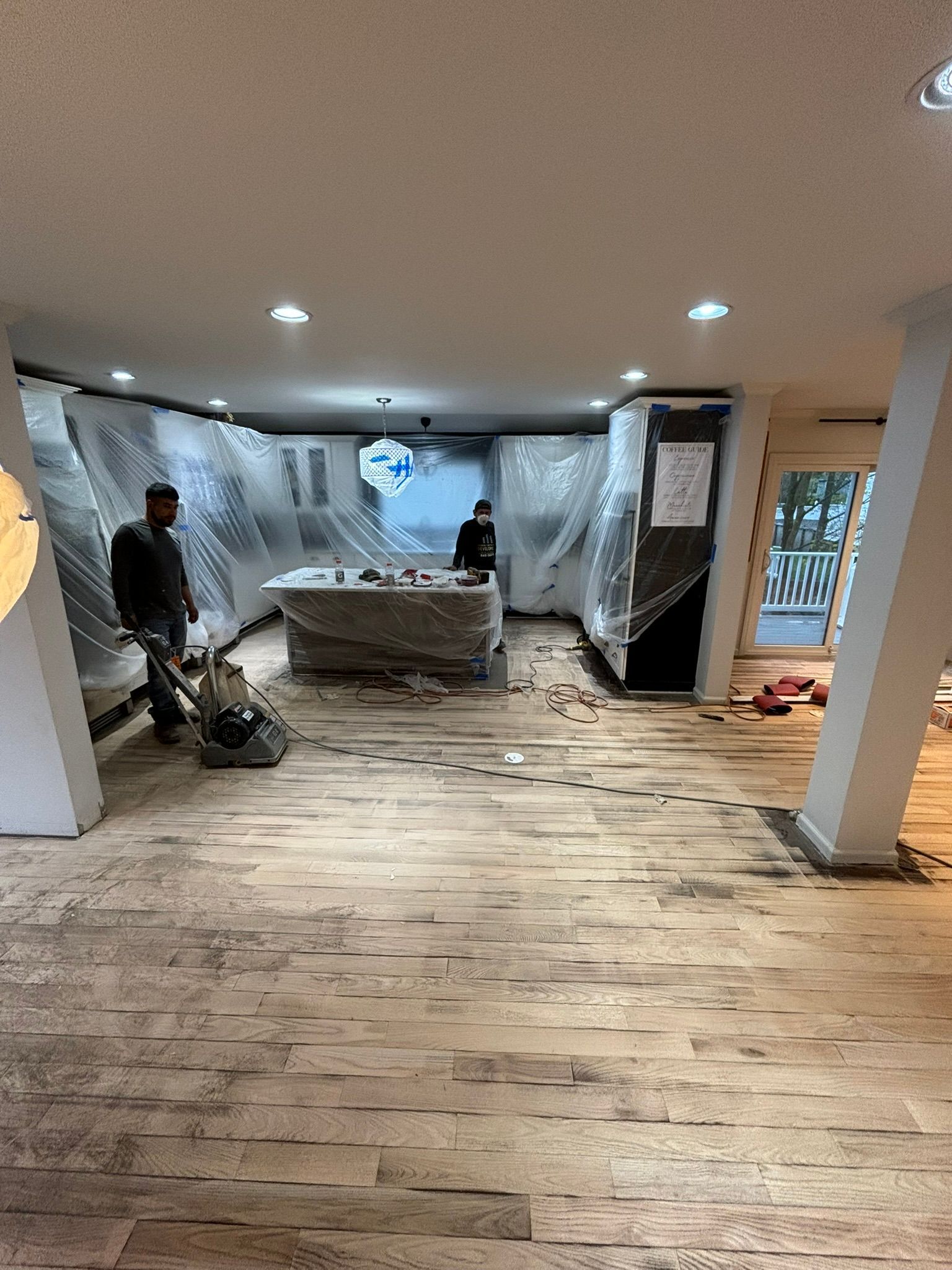 Men sanding a hardwood floor in a room under renovation, plastic sheeting covers walls and cabinets.