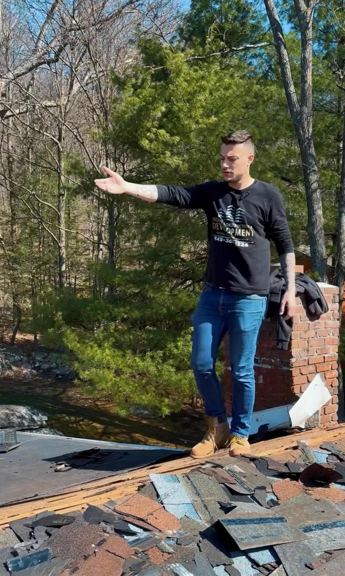 Man in jeans and black shirt on a roof, pointing to the left. Brick chimney and debris visible. Outdoors.
