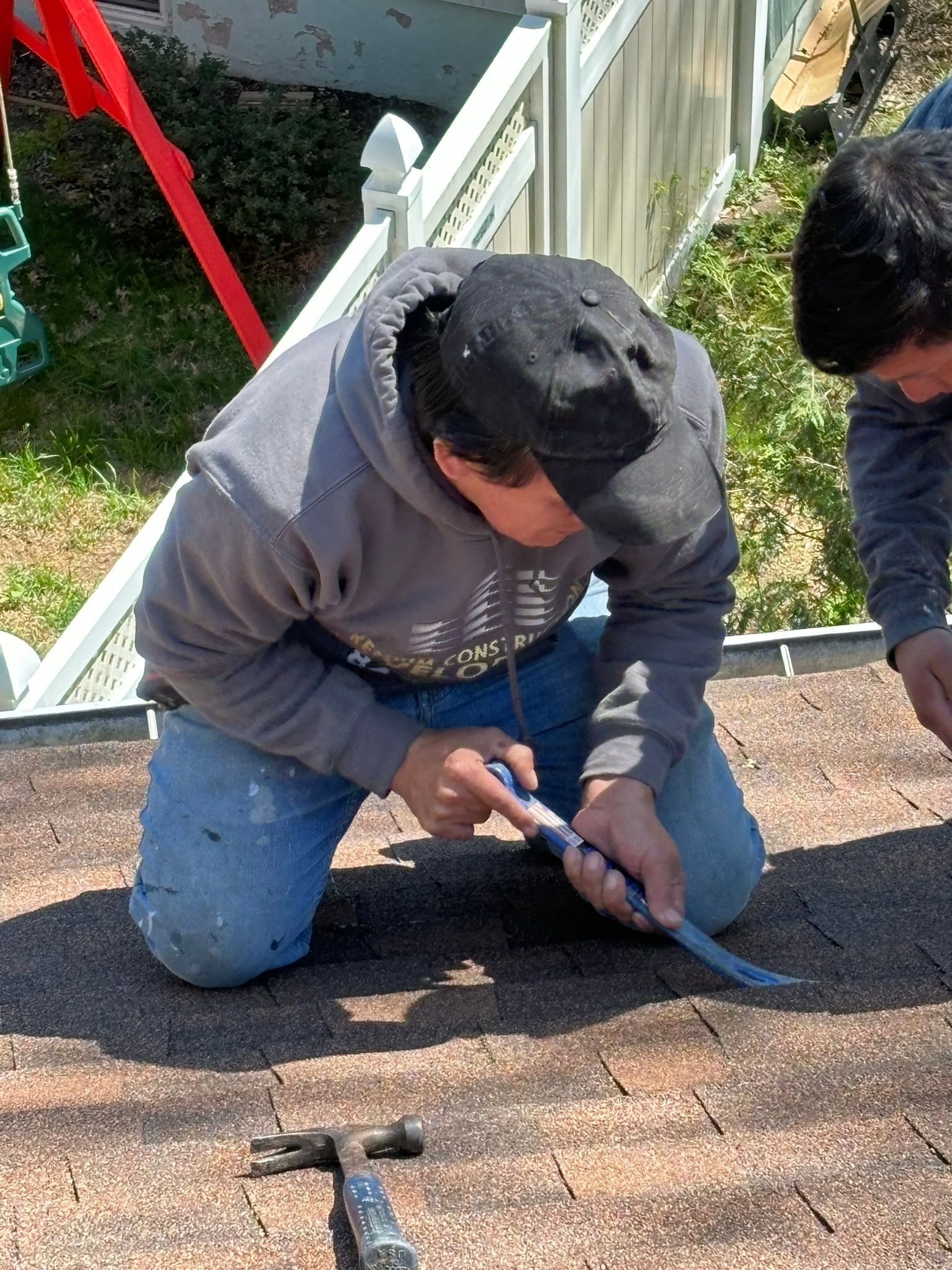 Man kneeling on a roof, removing shingles with a pry bar. He wears a gray hoodie, blue jeans, and a black cap.