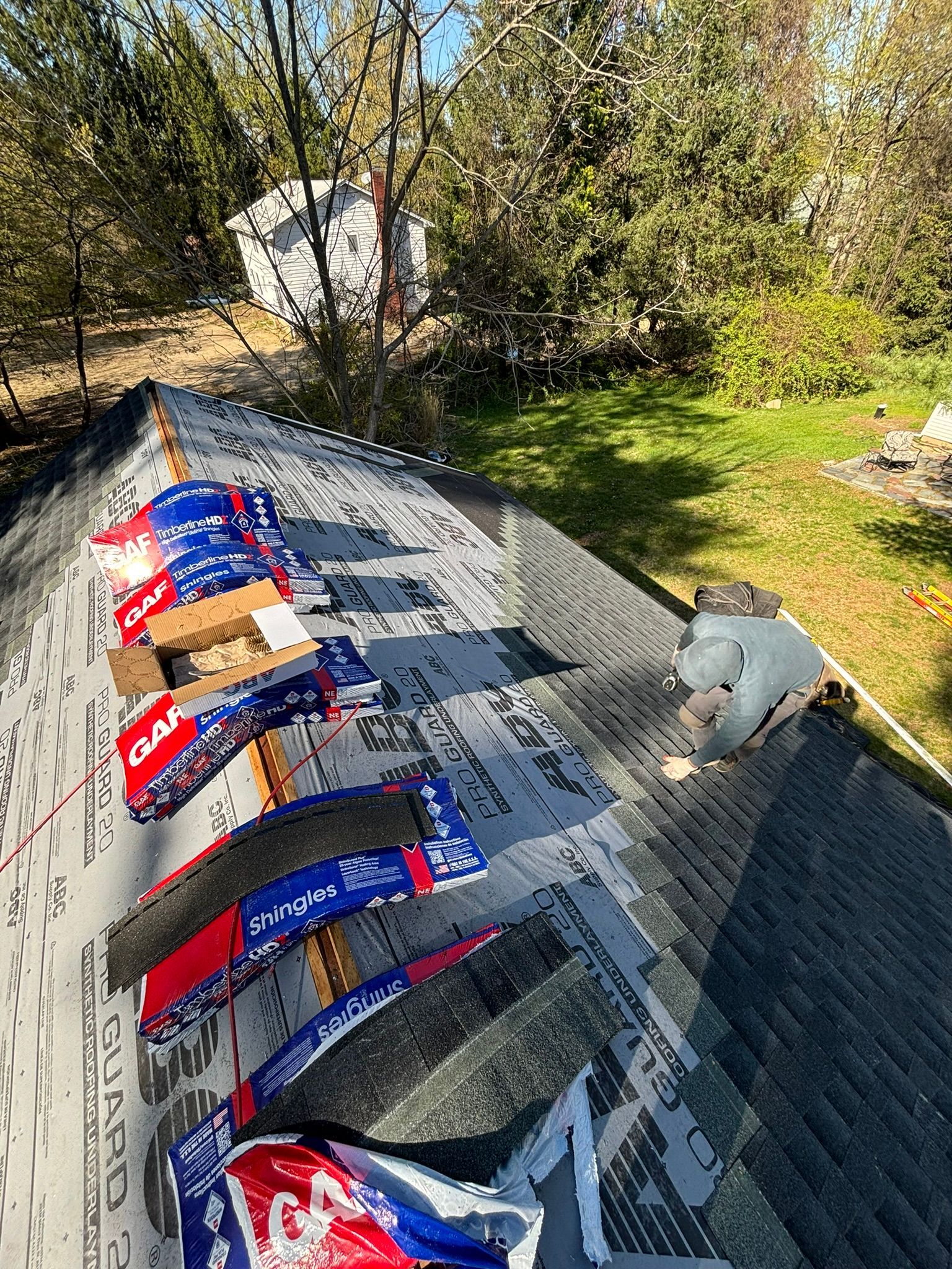 Roofing materials on a house roof, bags of shingles are in the foreground with a yard and trees in the background.