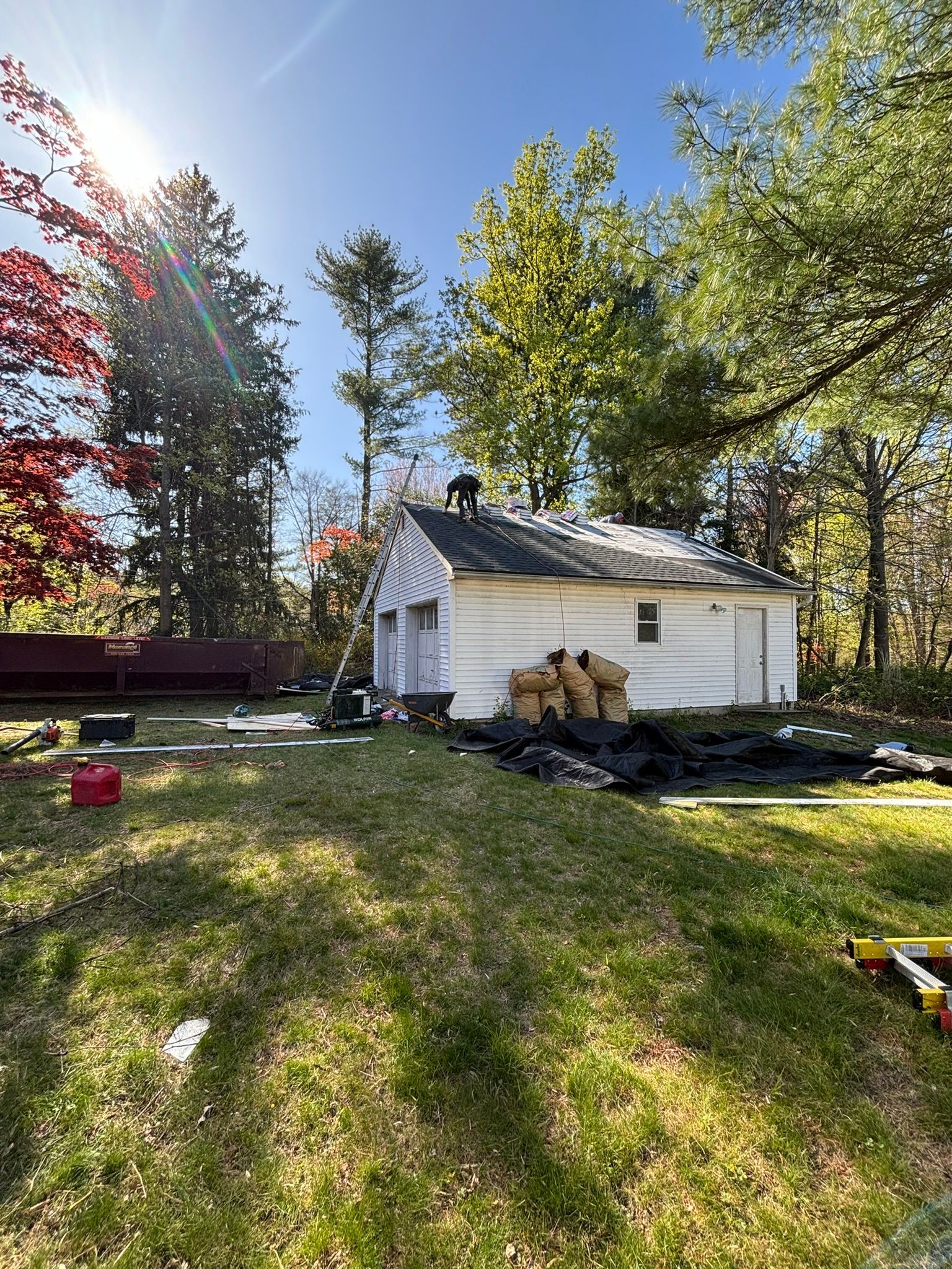 Garage with roof damage, surrounded by debris and trees on a sunny day.