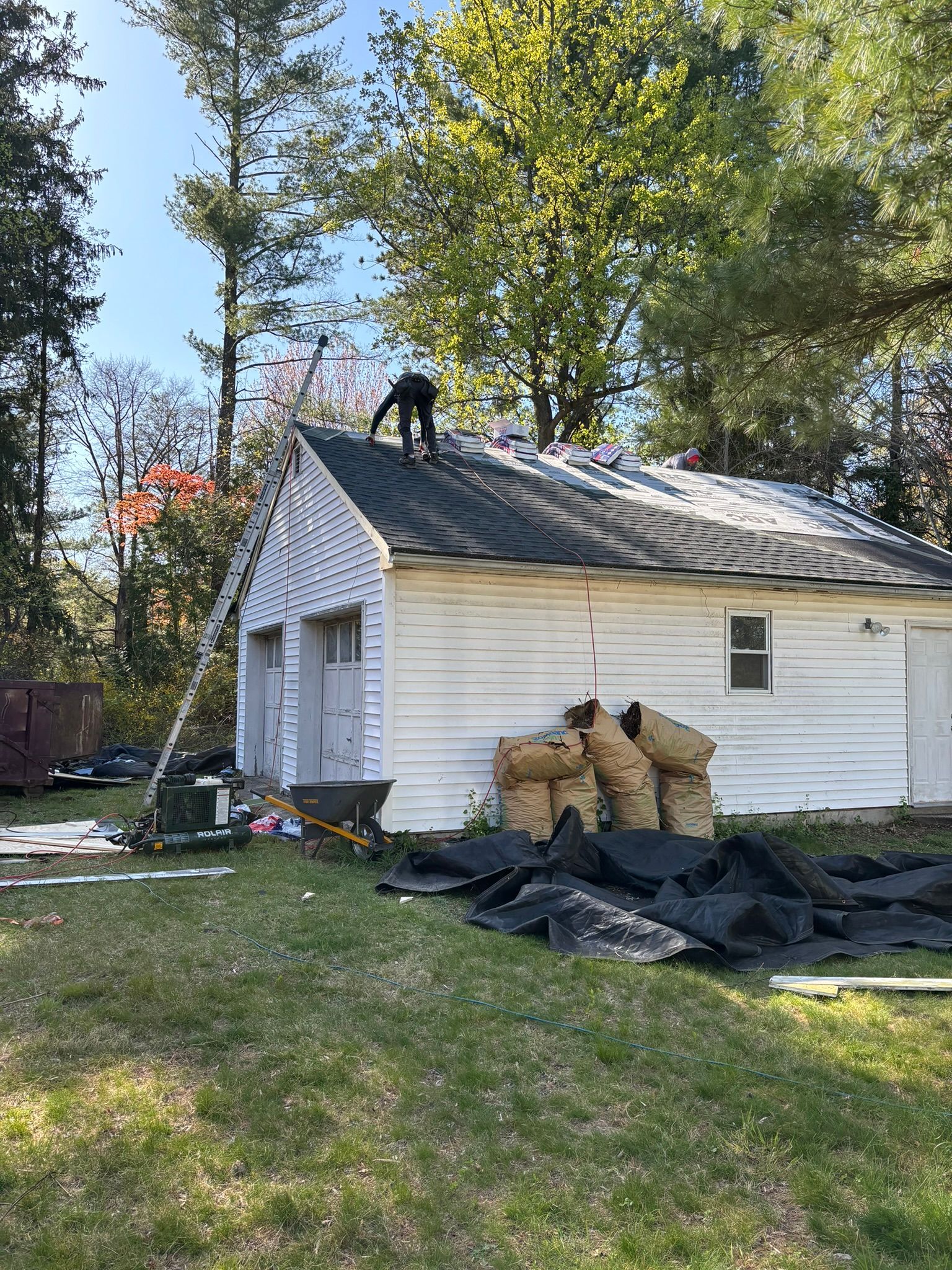 A person on a roof removing shingles from a white garage, with a ladder and debris on the ground.