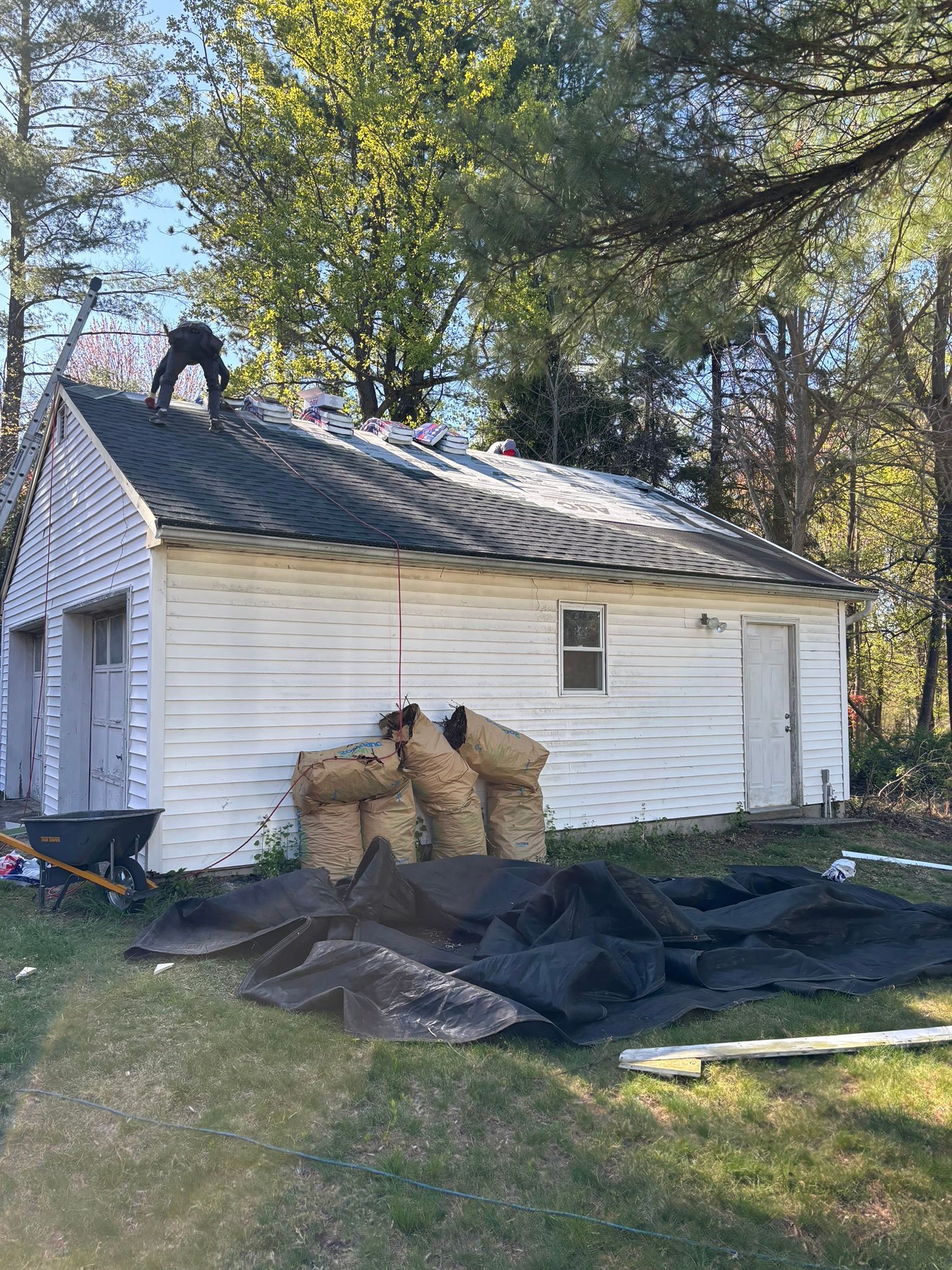 A worker on a white building's roof, replacing it. Brown bags and black tarps lie on the ground.