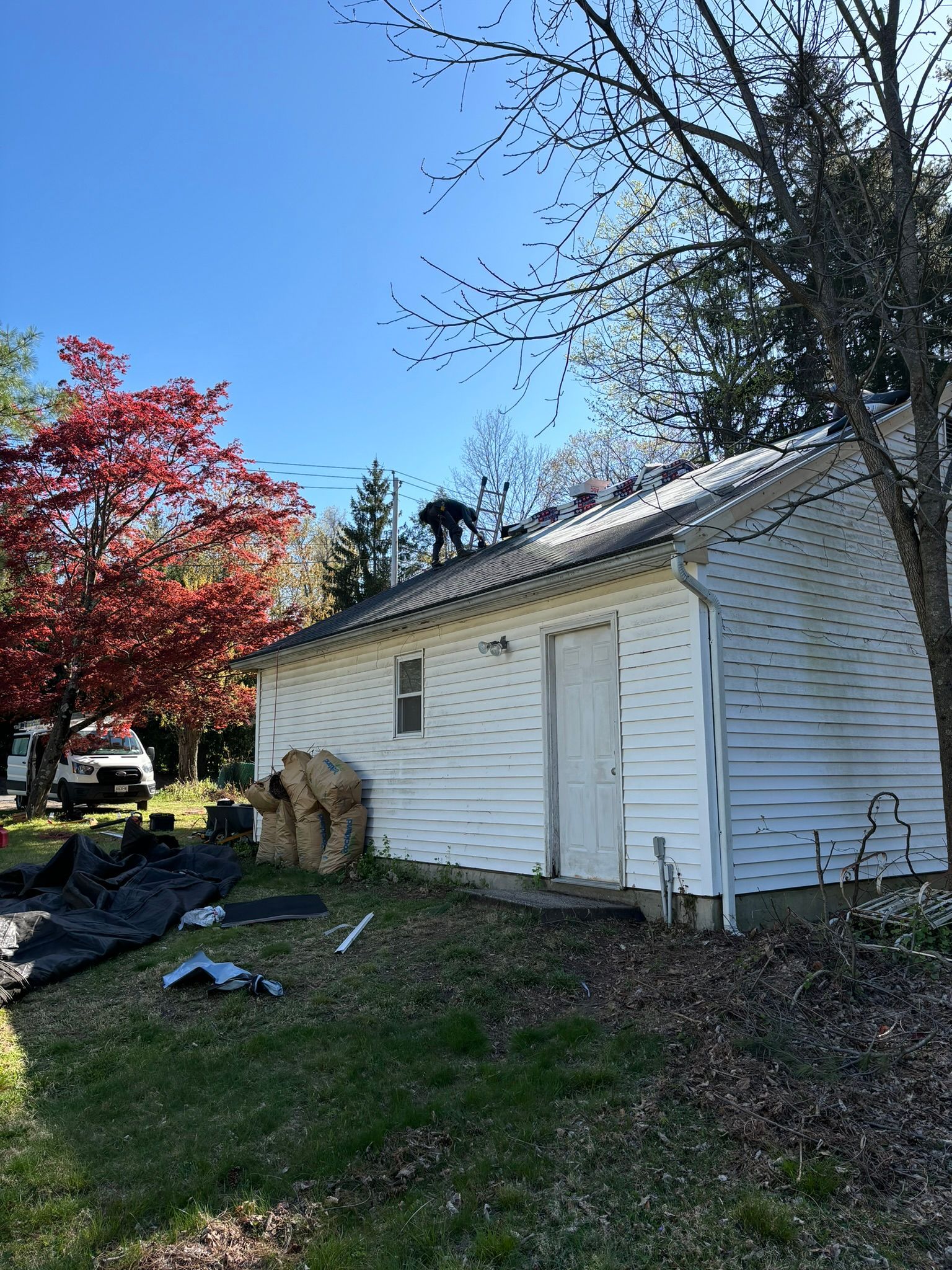 Roofer on a white house roof; red tree in background; black tarp and supplies in yard.