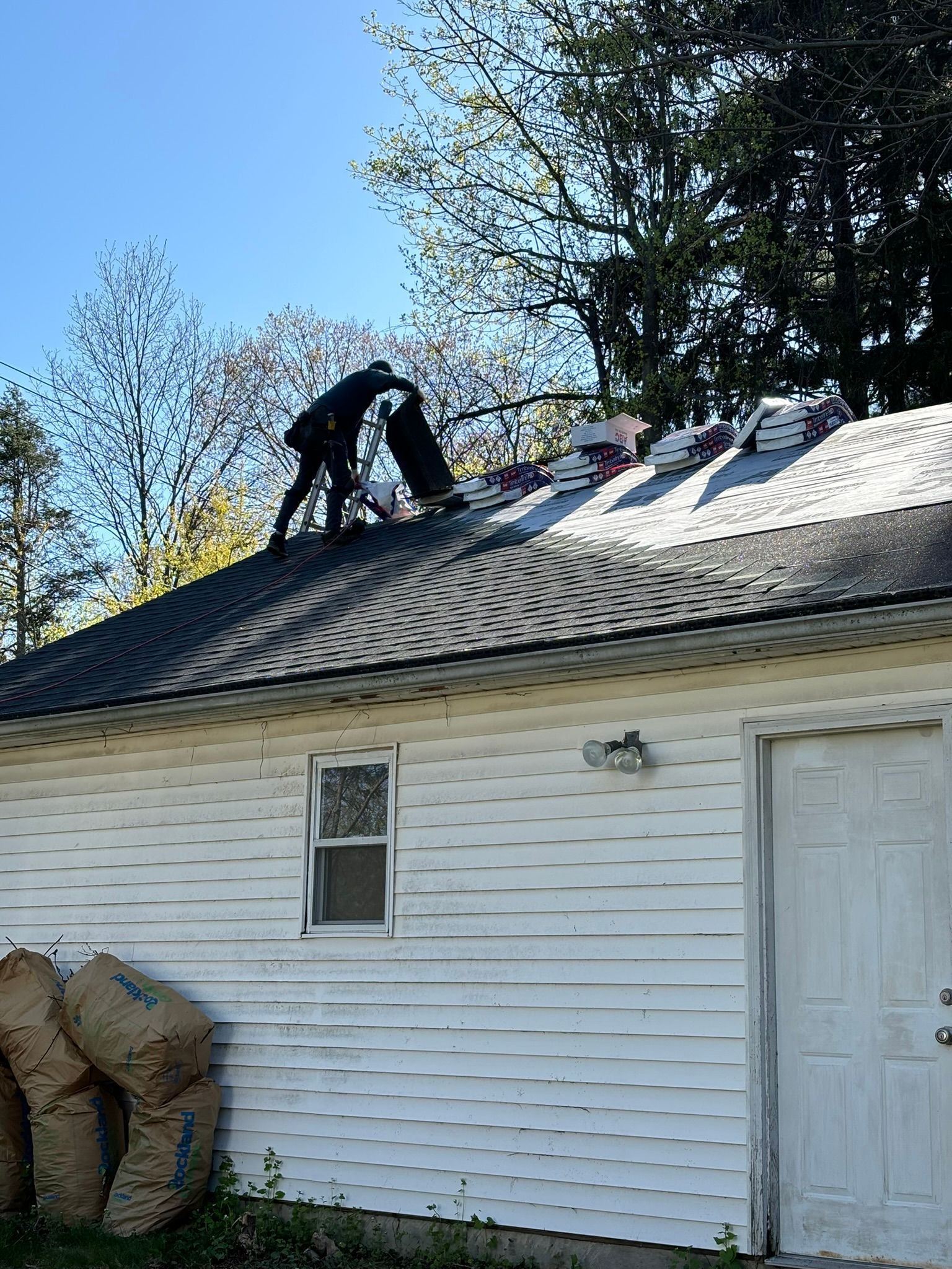 Roofer on a white-sided building, installing shingles on a rooftop under a blue sky.
