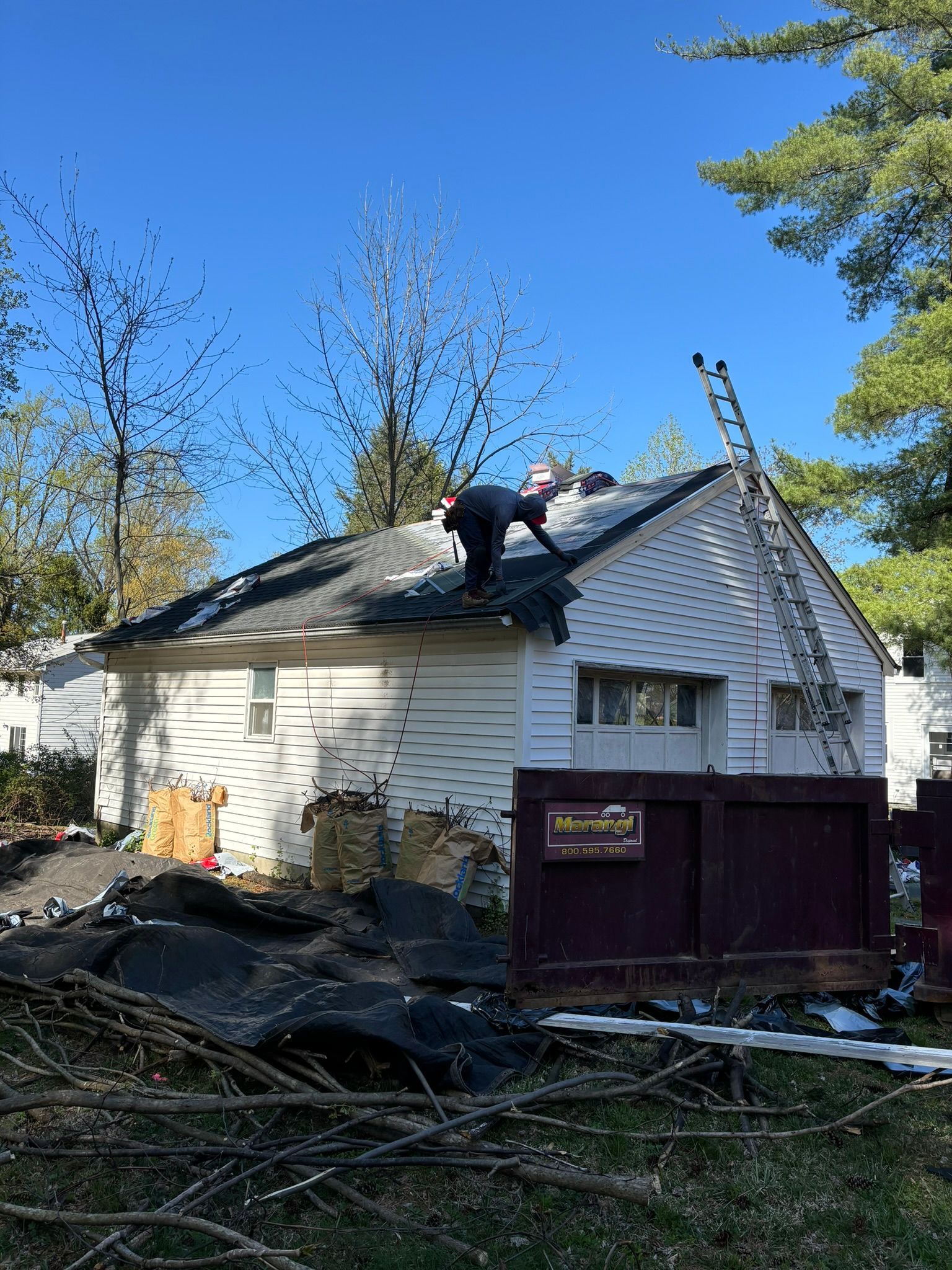 A roofer works on a white house roof under a clear blue sky. A ladder leans against the house.