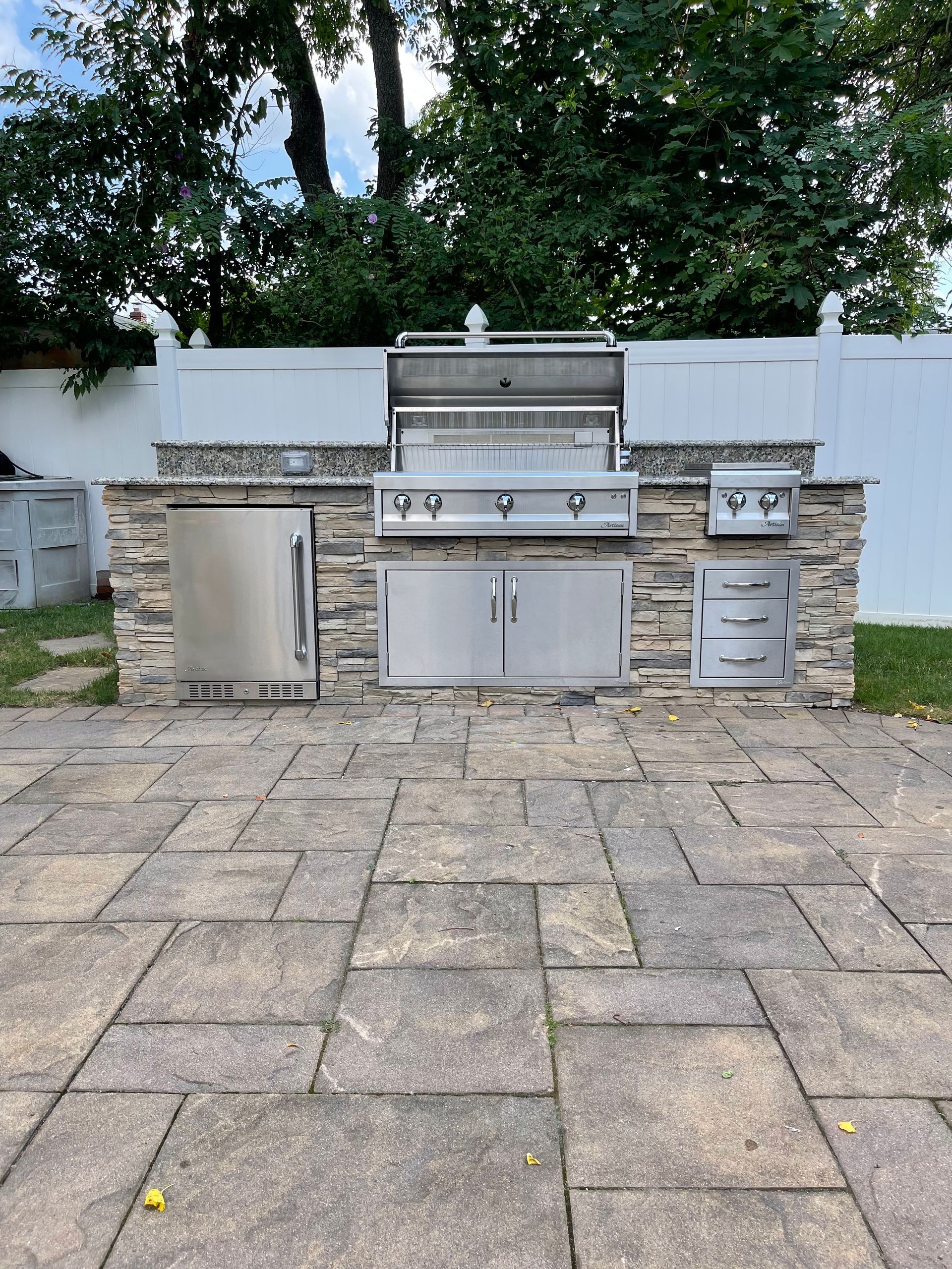 Outdoor kitchen with stone facade, stainless steel grill, refrigerator, and storage on a brick patio.