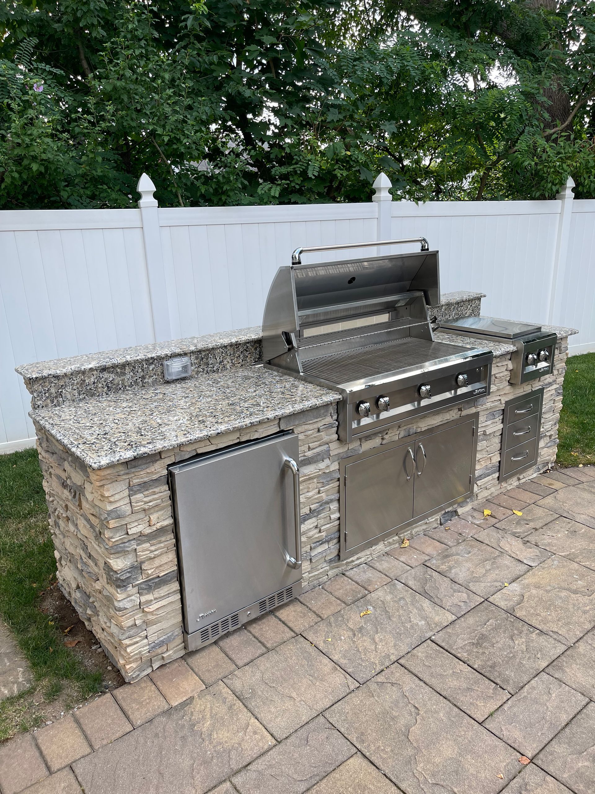 Outdoor kitchen with a grill, fridge, and stone facade on a brick patio.