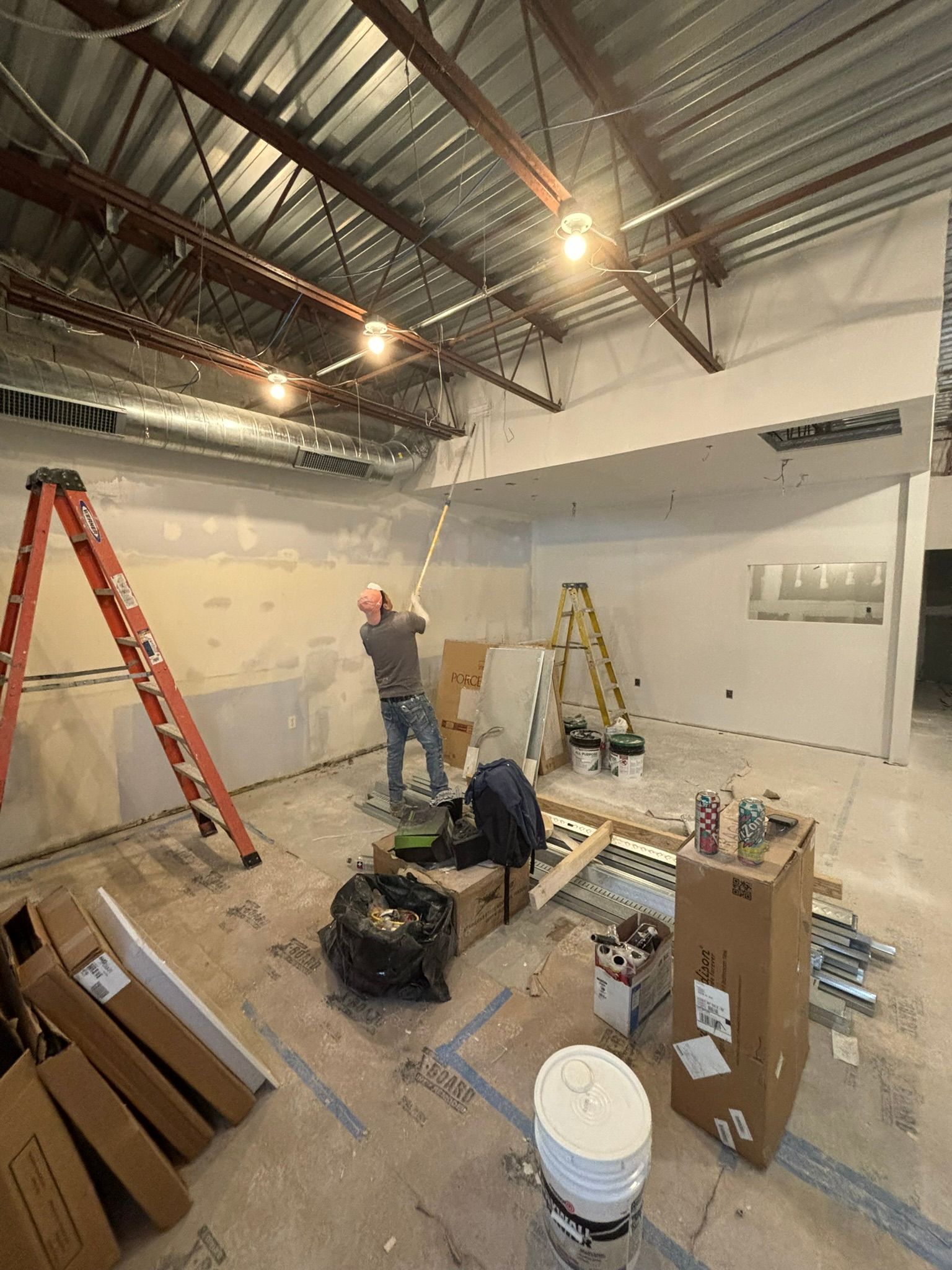Construction site: man on ladder working on ceiling. Tools and materials scattered; orange and yellow ladders visible.