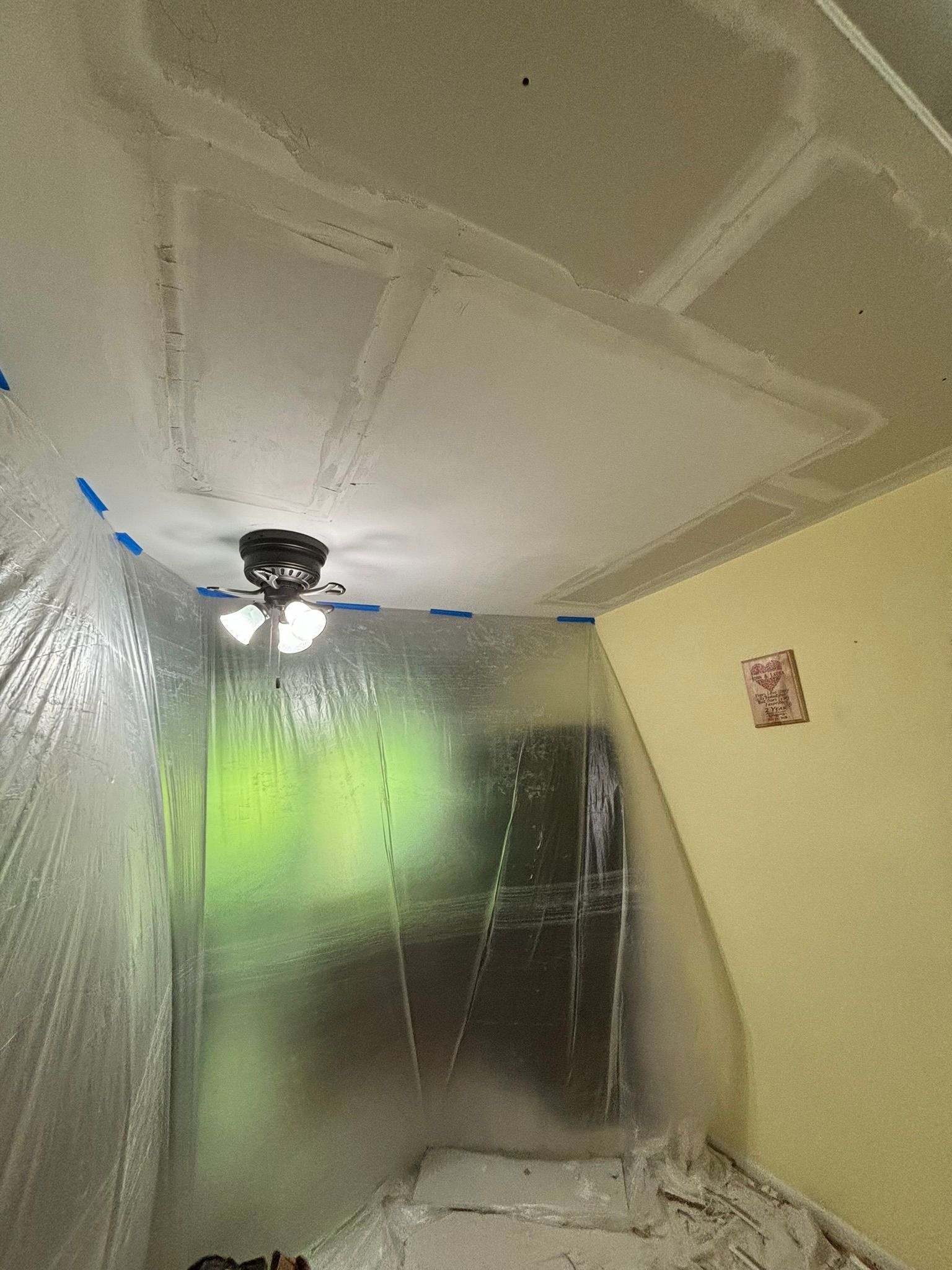 Room with ceiling repair in progress. Plastic sheeting protects walls; ceiling shows patched drywall, tape, and light.