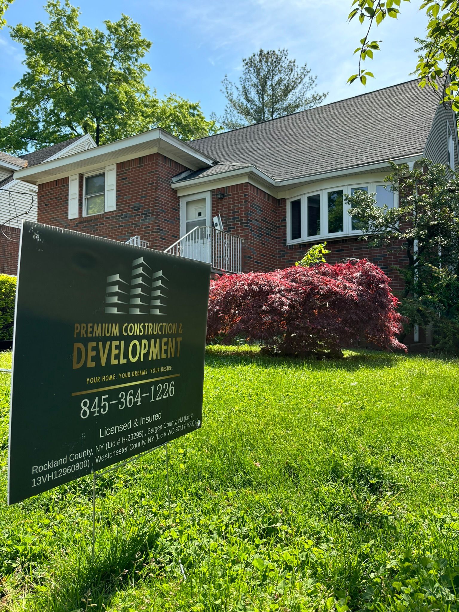 Brick house with development sign on front lawn, under blue sky.