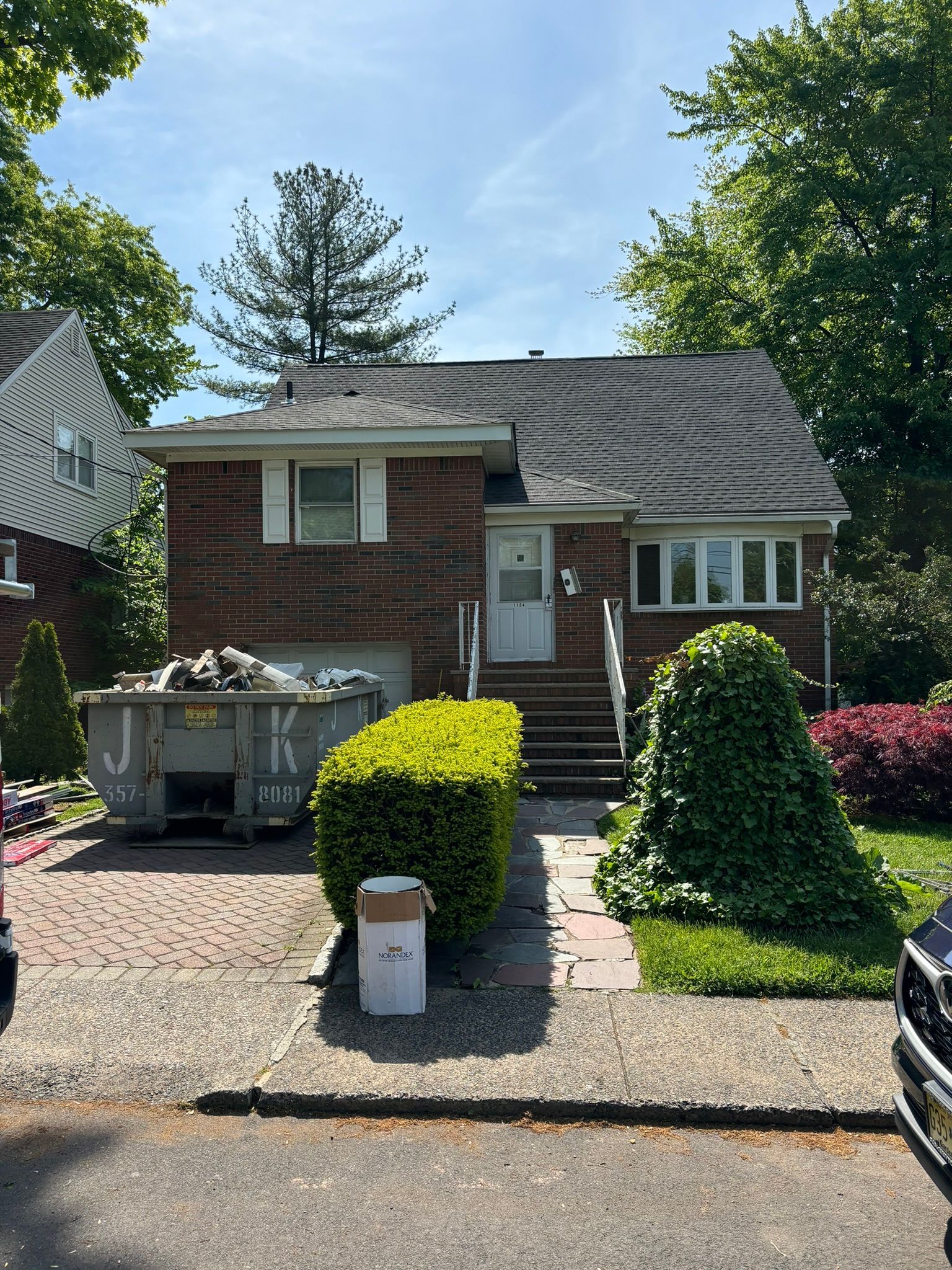 Brick house with dumpster in front; bushes and steps lead to the front door.