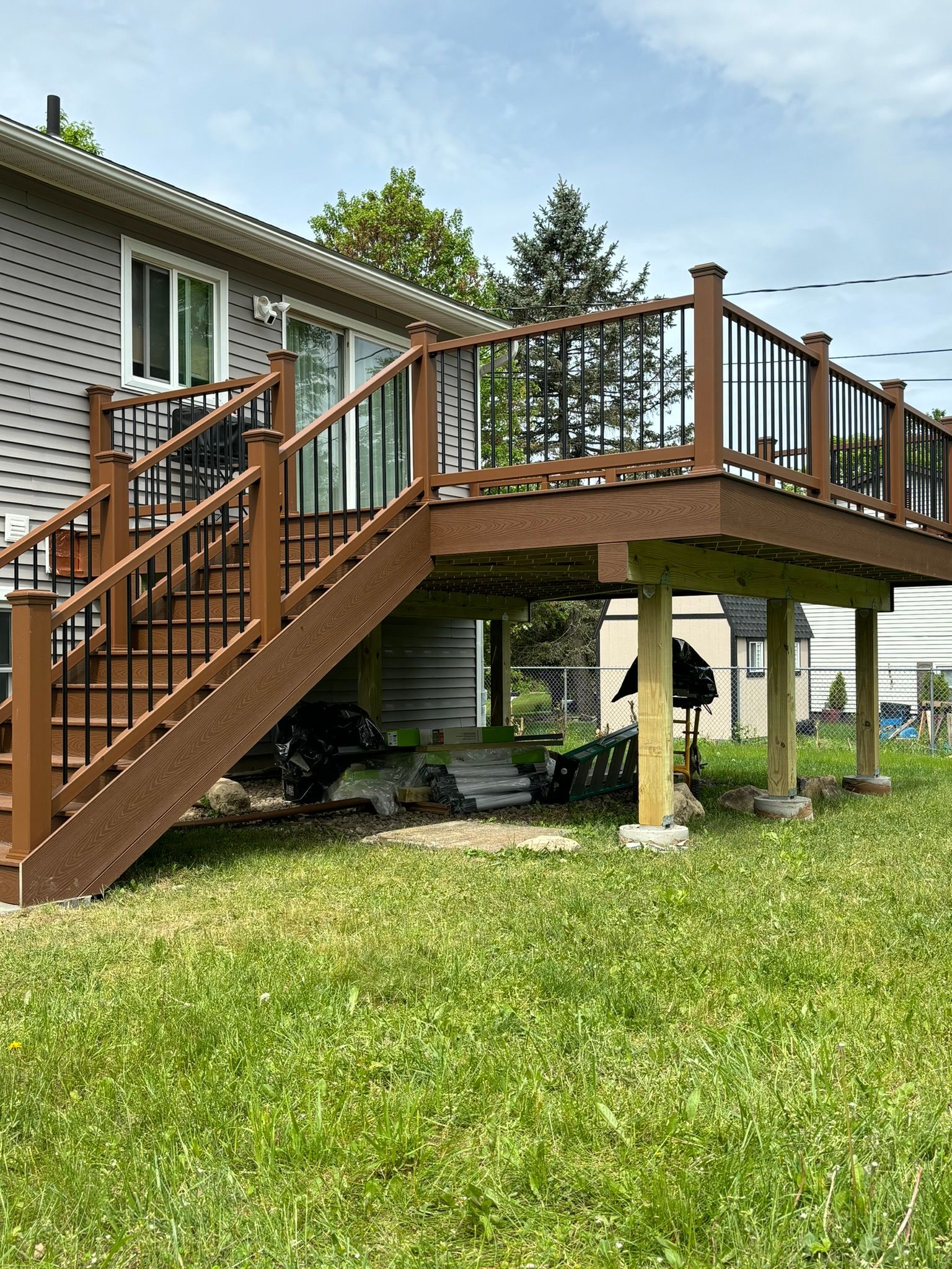 Brown composite deck with stairs attached to a gray house and supported by wooden posts in a grassy yard.