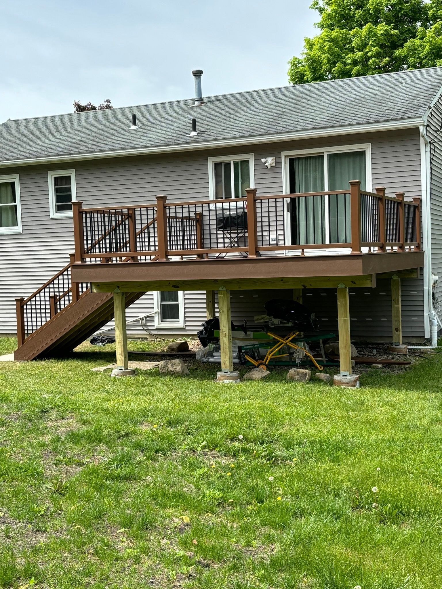 Brown deck attached to a gray house with a staircase, supported by wooden posts on concrete bases.