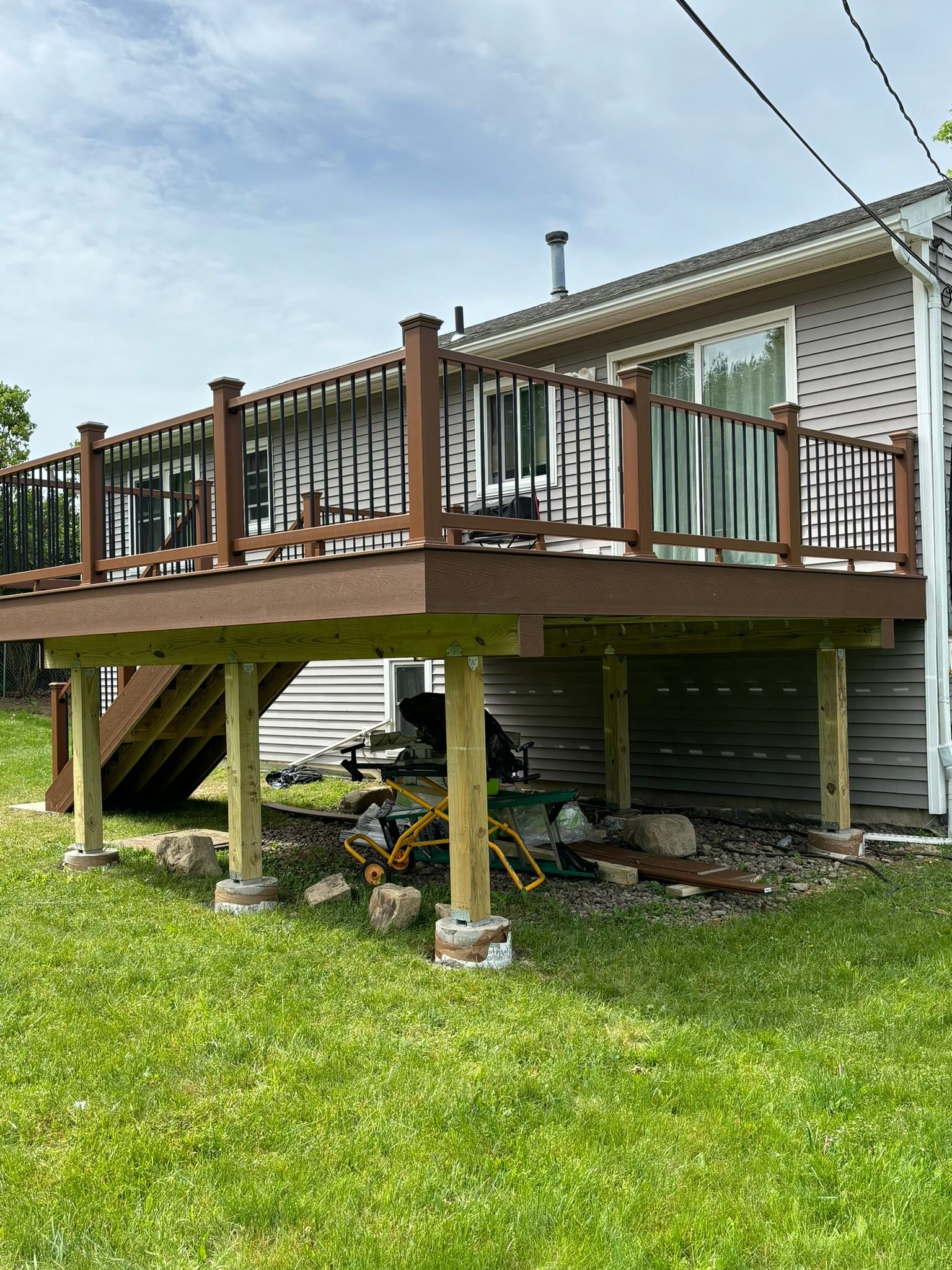 Elevated deck with brown composite boards, black railing, and wooden posts, attached to a house with siding.