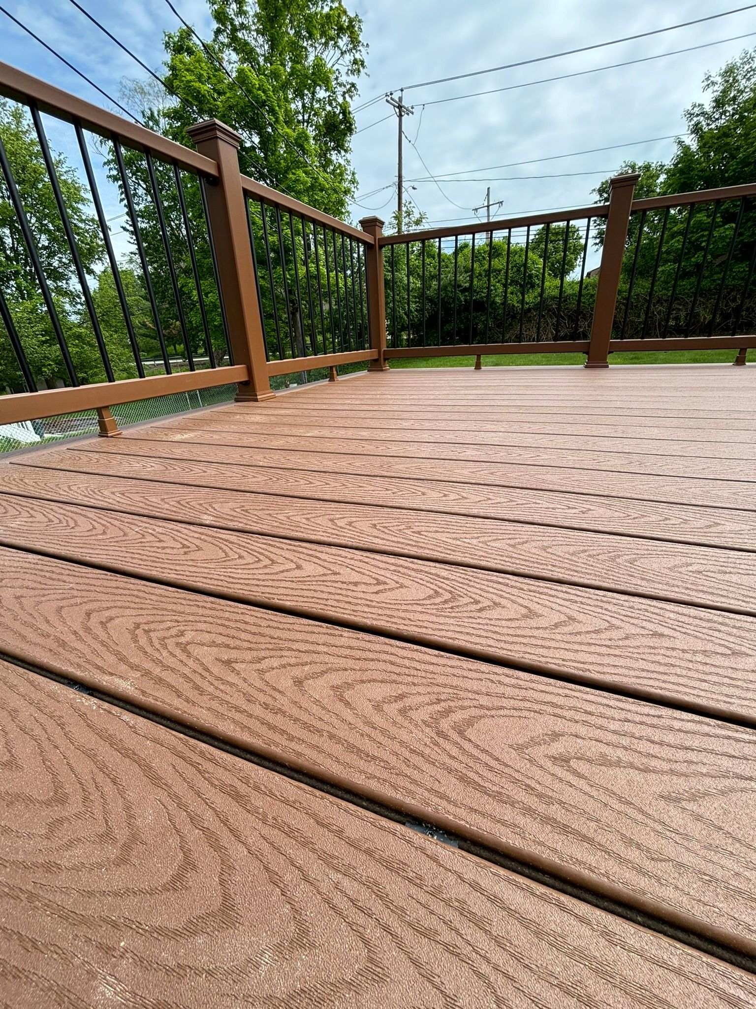 Brown composite deck with black railing, against green trees and blue sky.