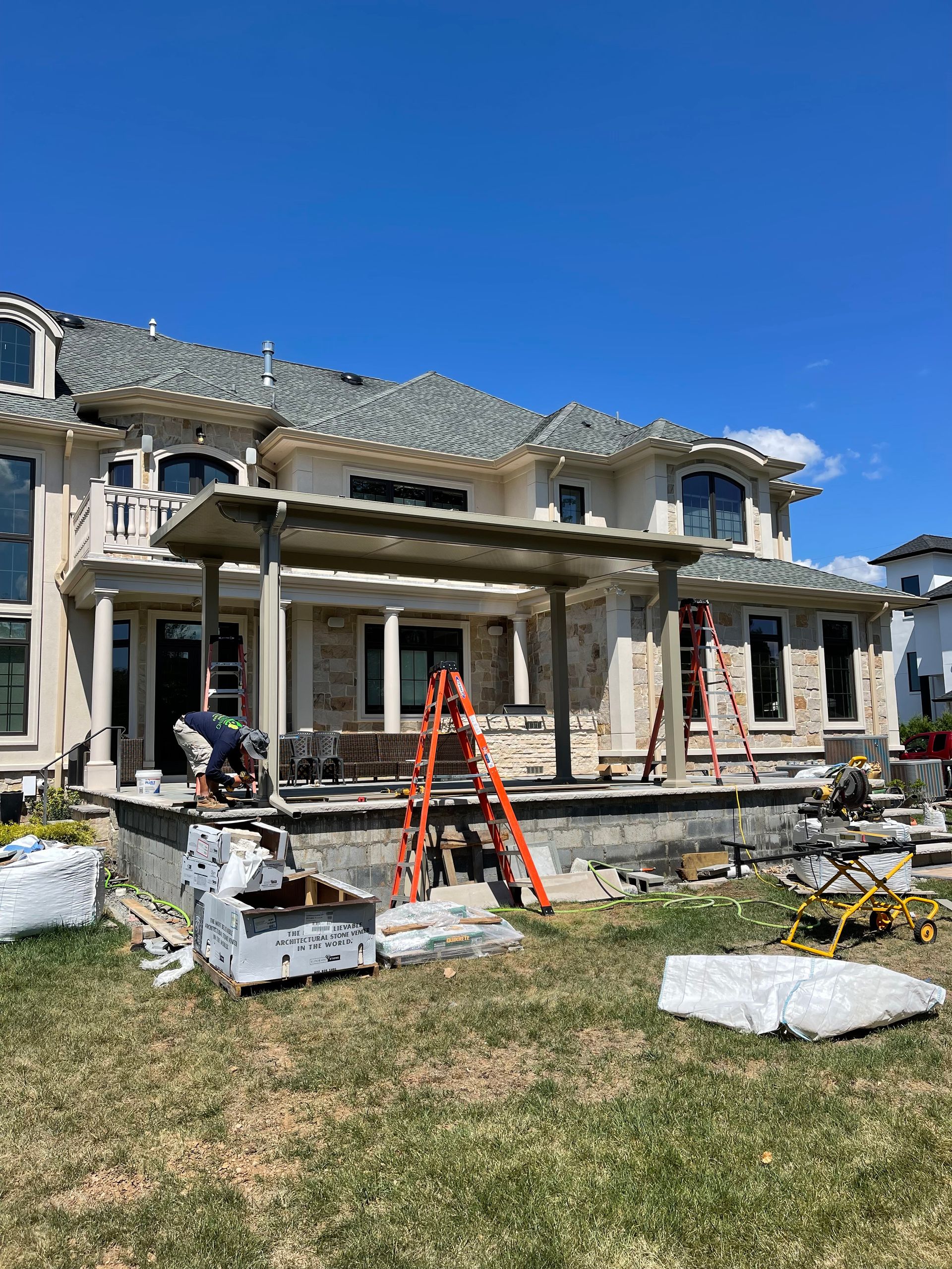 Backyard patio construction at a large house; workers, ladders, materials scattered on the grass.