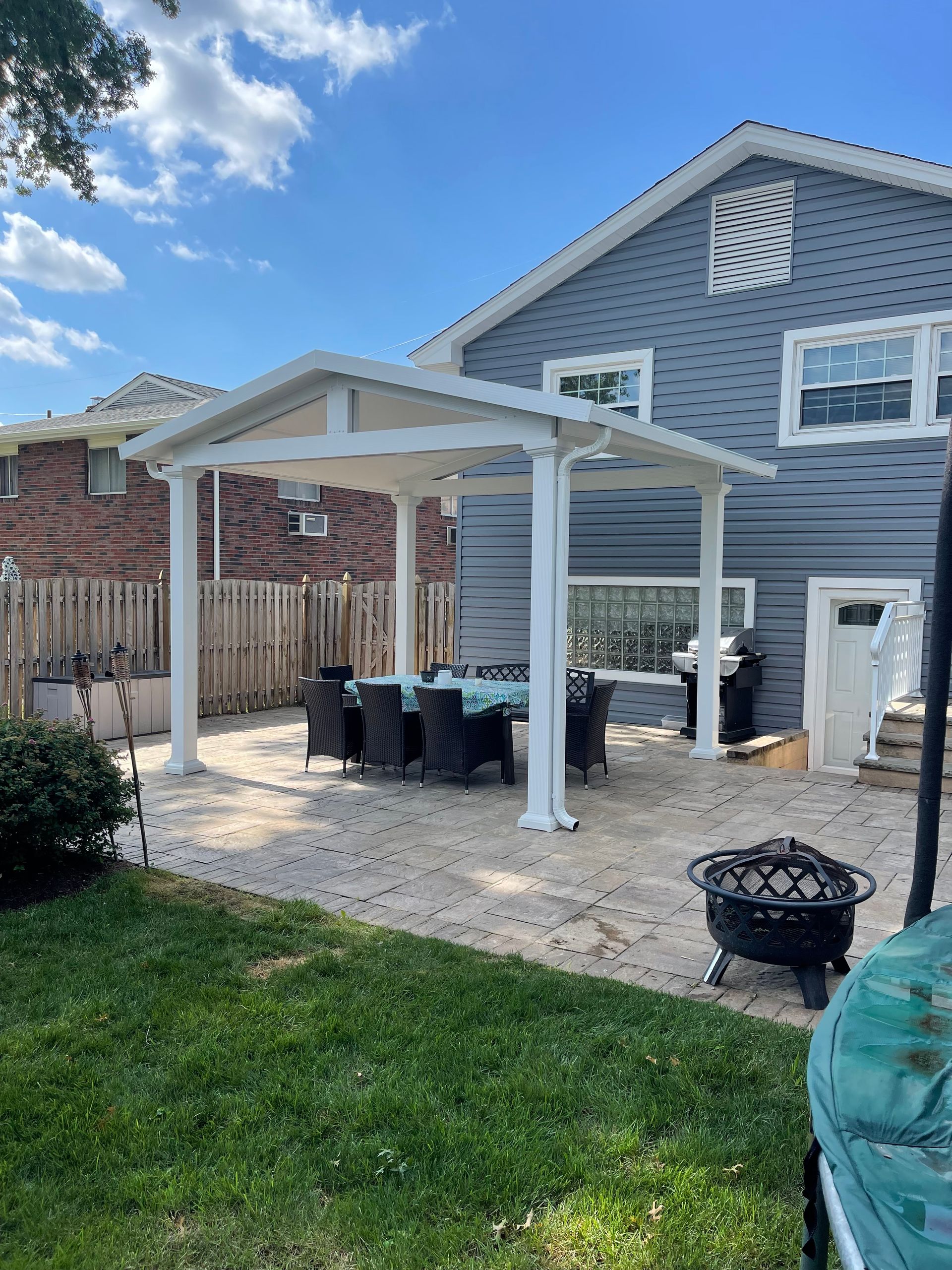 Backyard patio with white pergola, dining table, and grill next to a gray house.