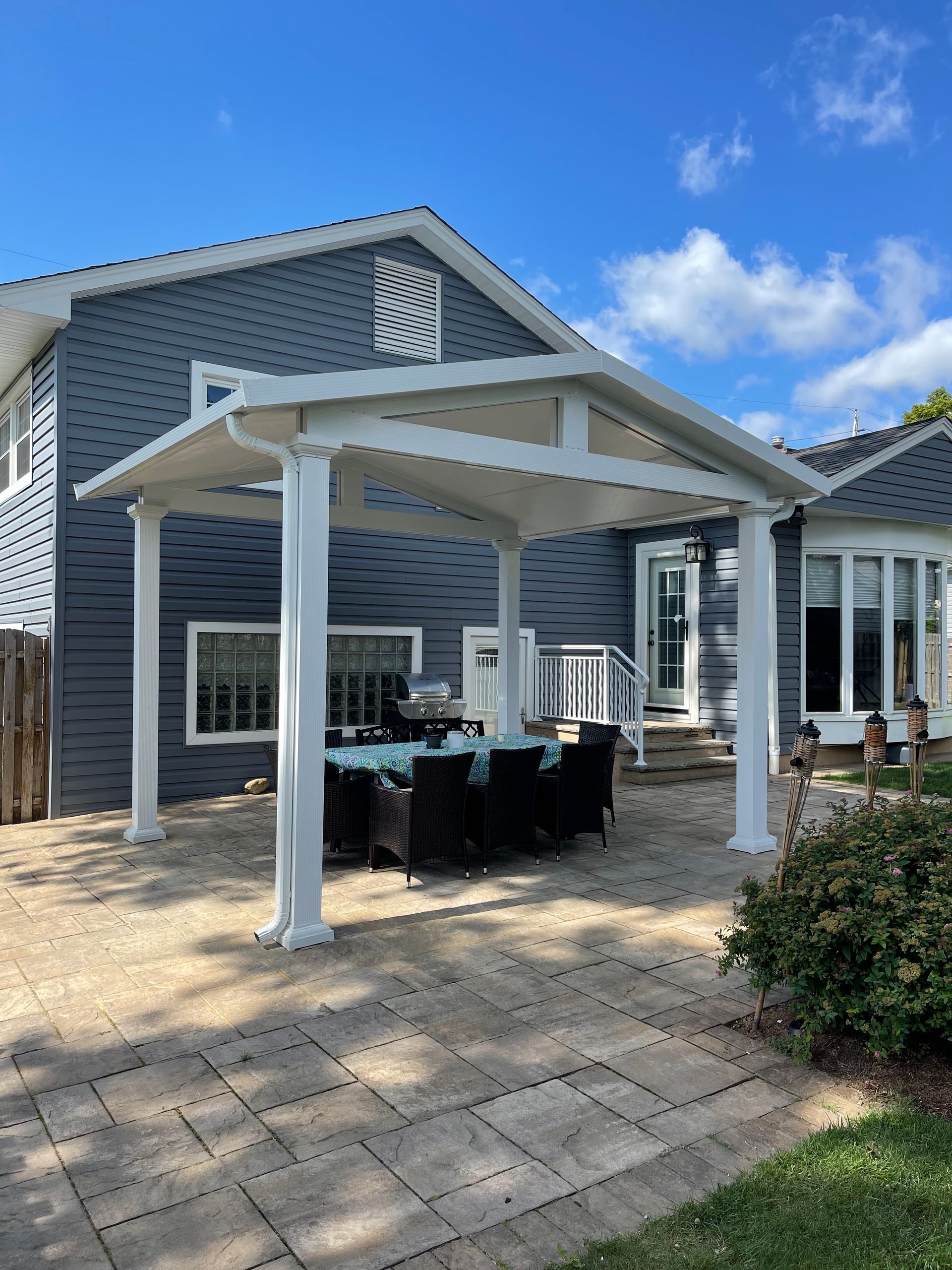 White pergola over patio with dining table. Gray siding on the house, blue sky overhead.