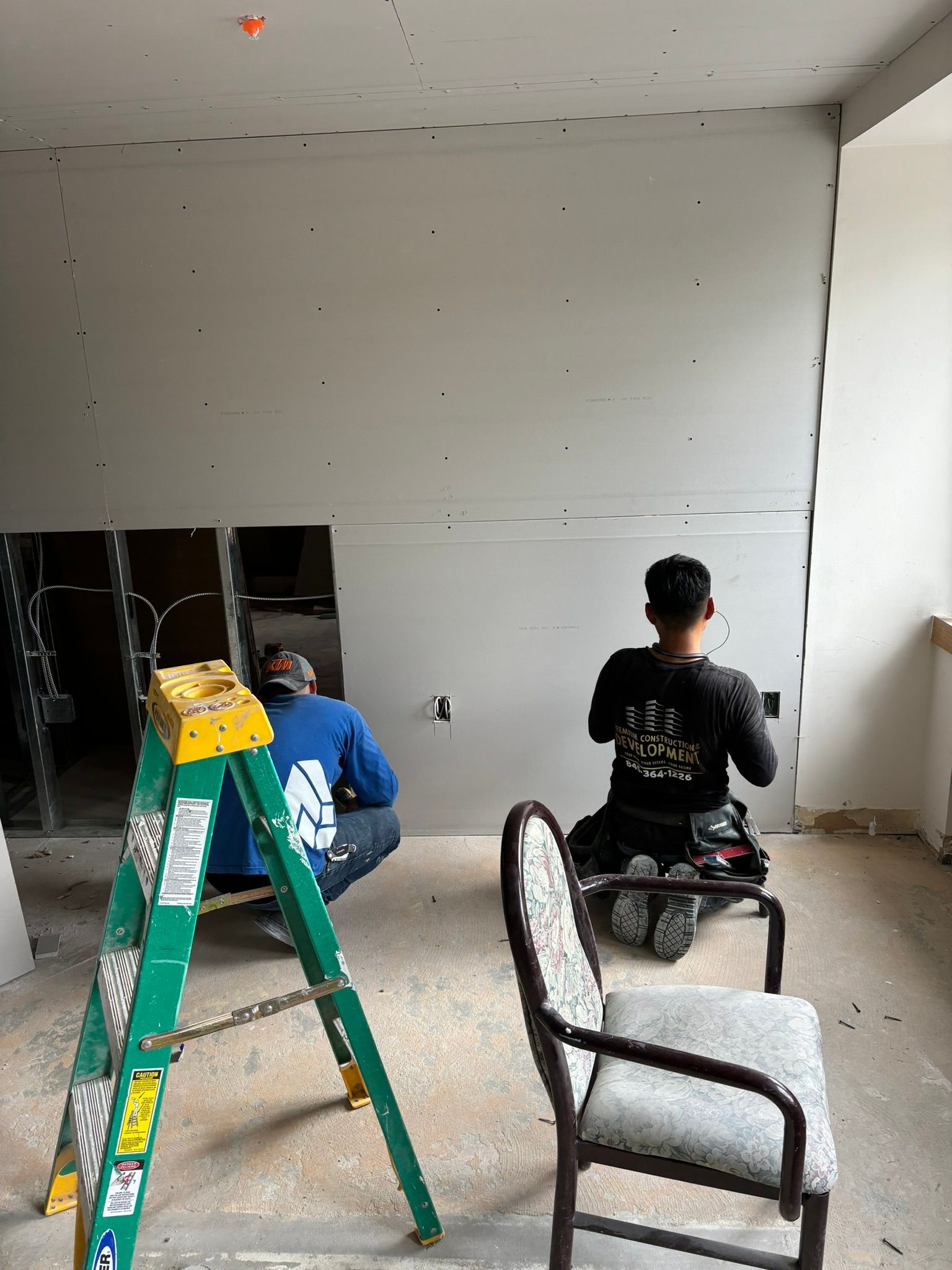 Two workers installing drywall on a wall. One kneels, the other squats. A ladder and chair are present.