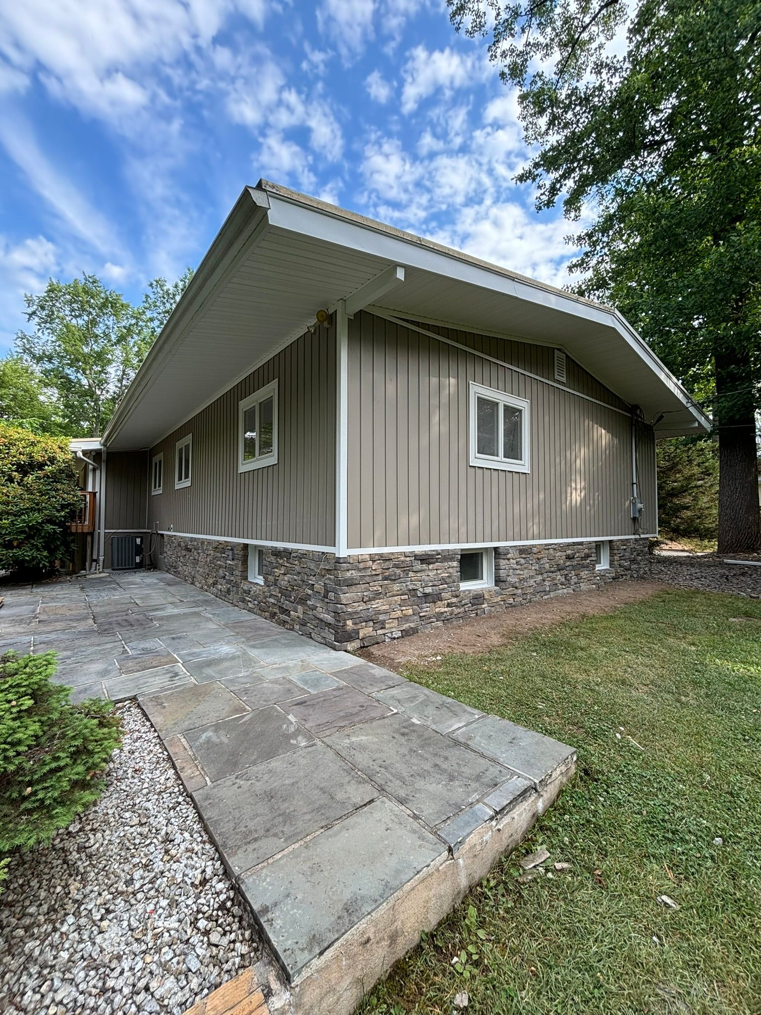 A house with a stone base, tan siding, and a sloped roof; a gray walkway leads to the door.