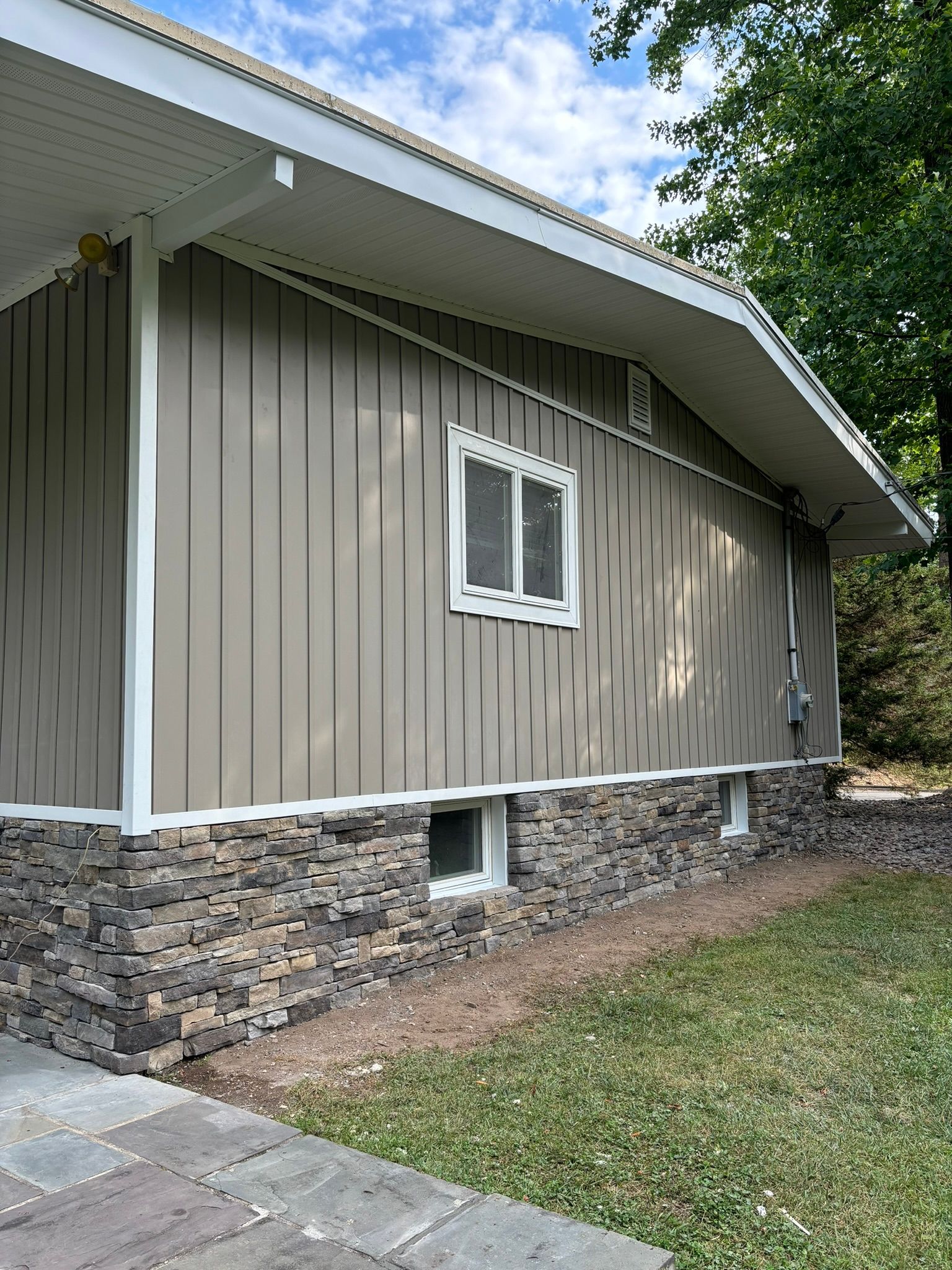 Tan vertical siding with stone veneer foundation, white trim, and window.