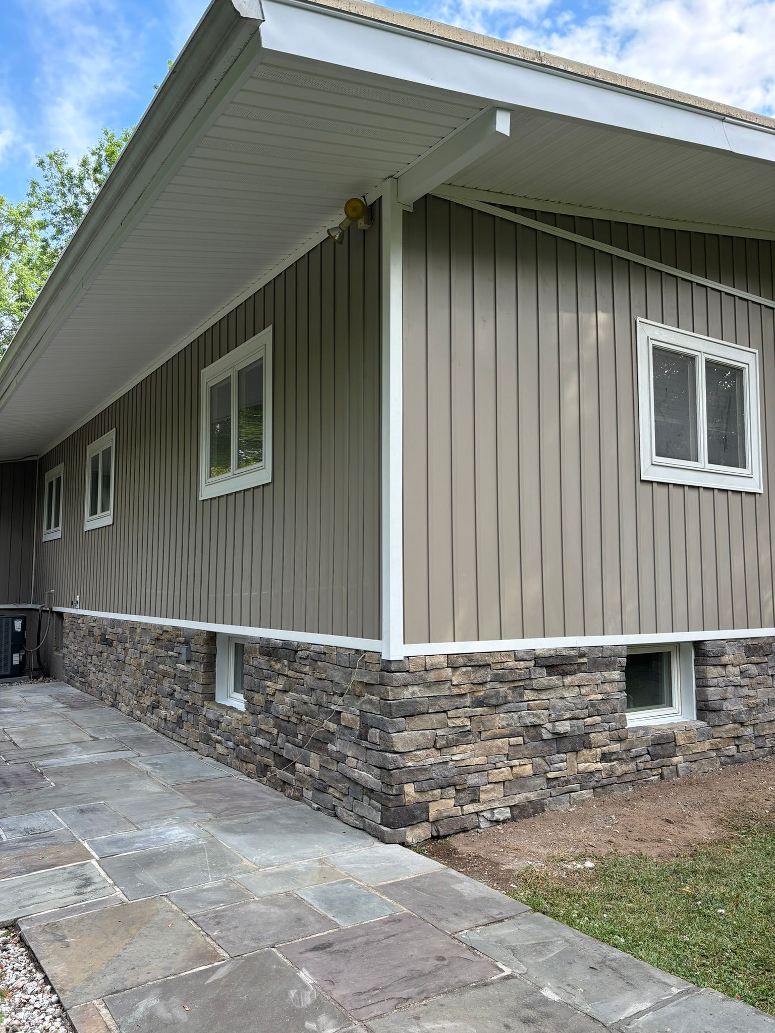 Beige house with stone base, white trim, and windows; paved walkway.