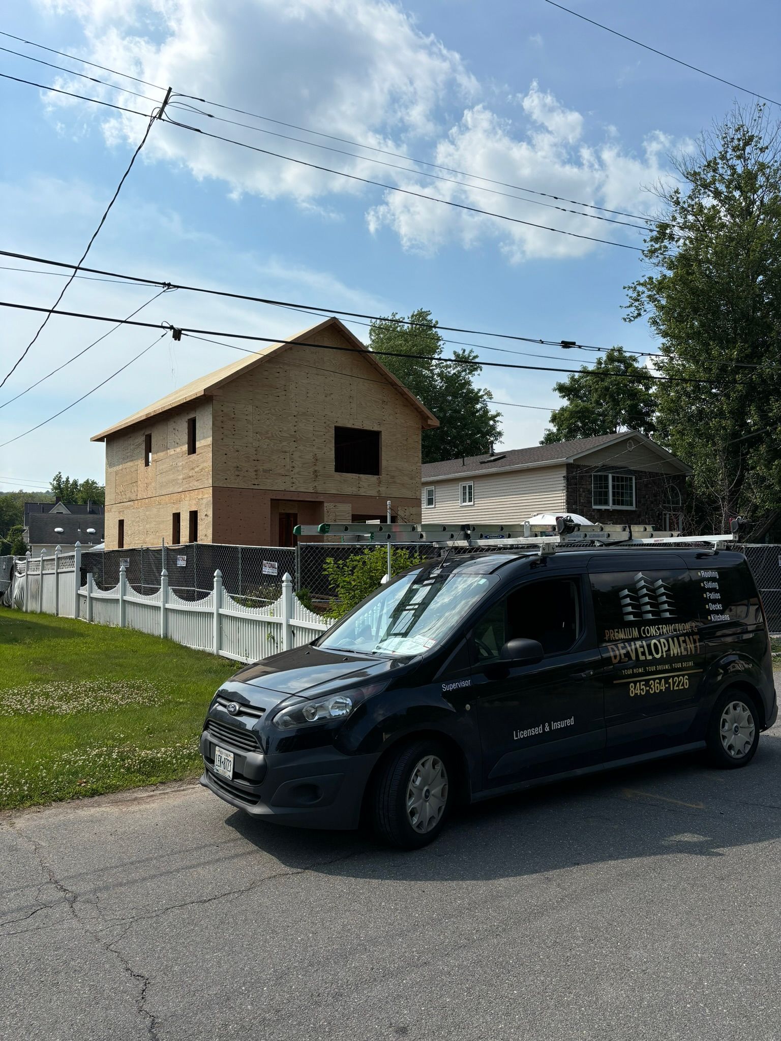Black van parked on a street; a house under construction and a white picket fence are in the background under a blue sky.
