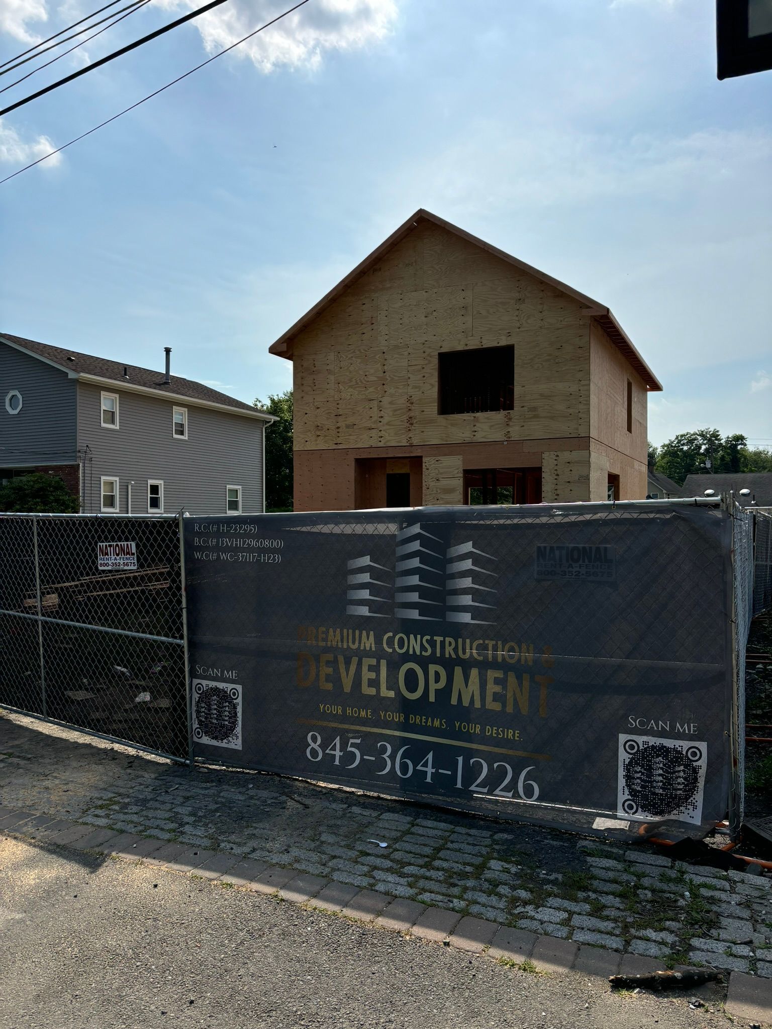 A new house under construction behind a construction fence; next to a finished house under a blue sky.