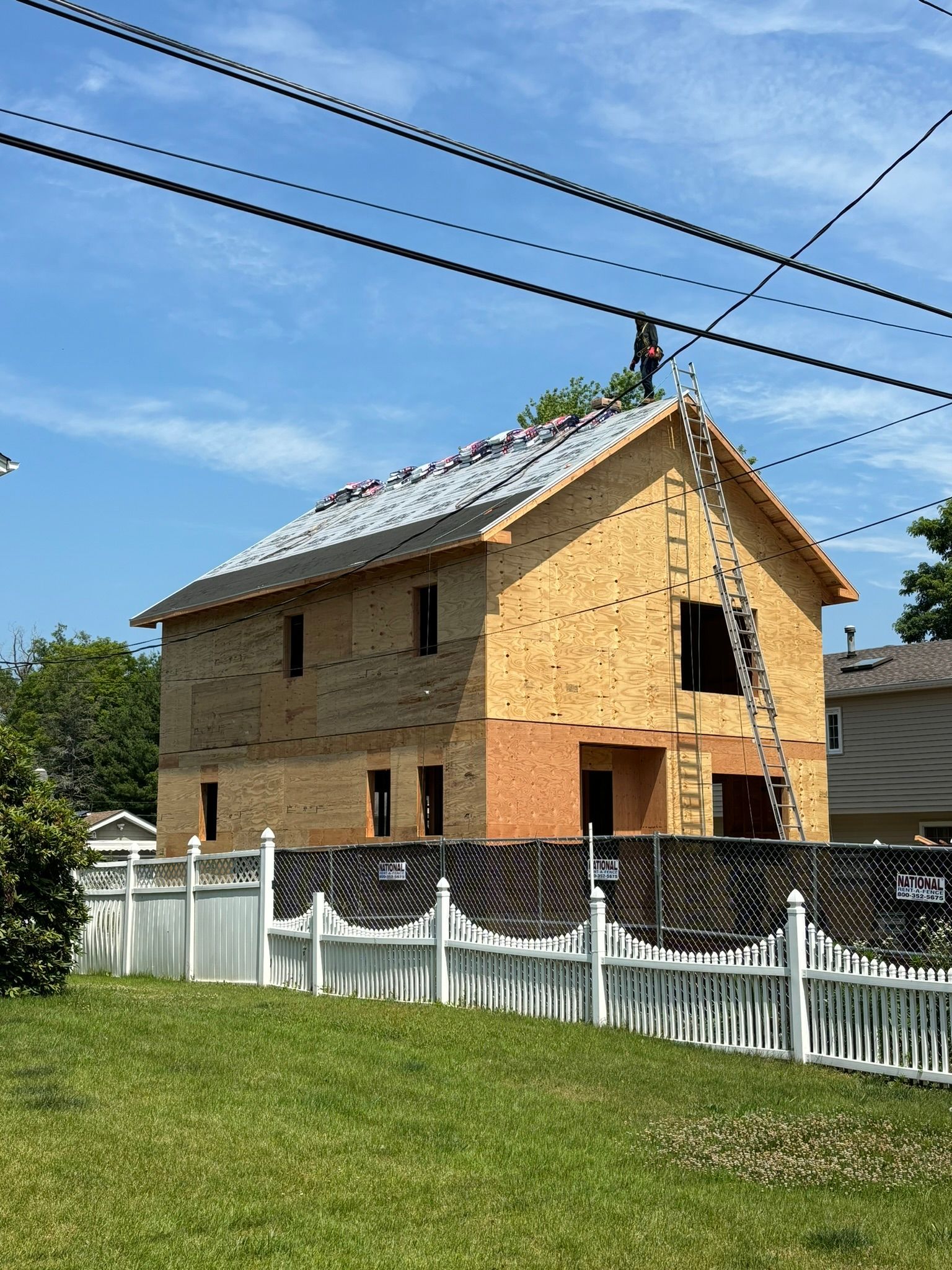 Two-story house under construction behind a white picket fence, surrounded by greenery and power lines.