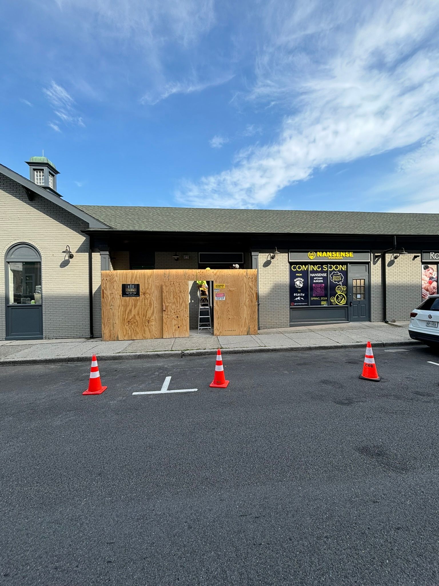 Exterior view of a building with a boarded-up entrance. Orange traffic cones mark a safety zone on the pavement.