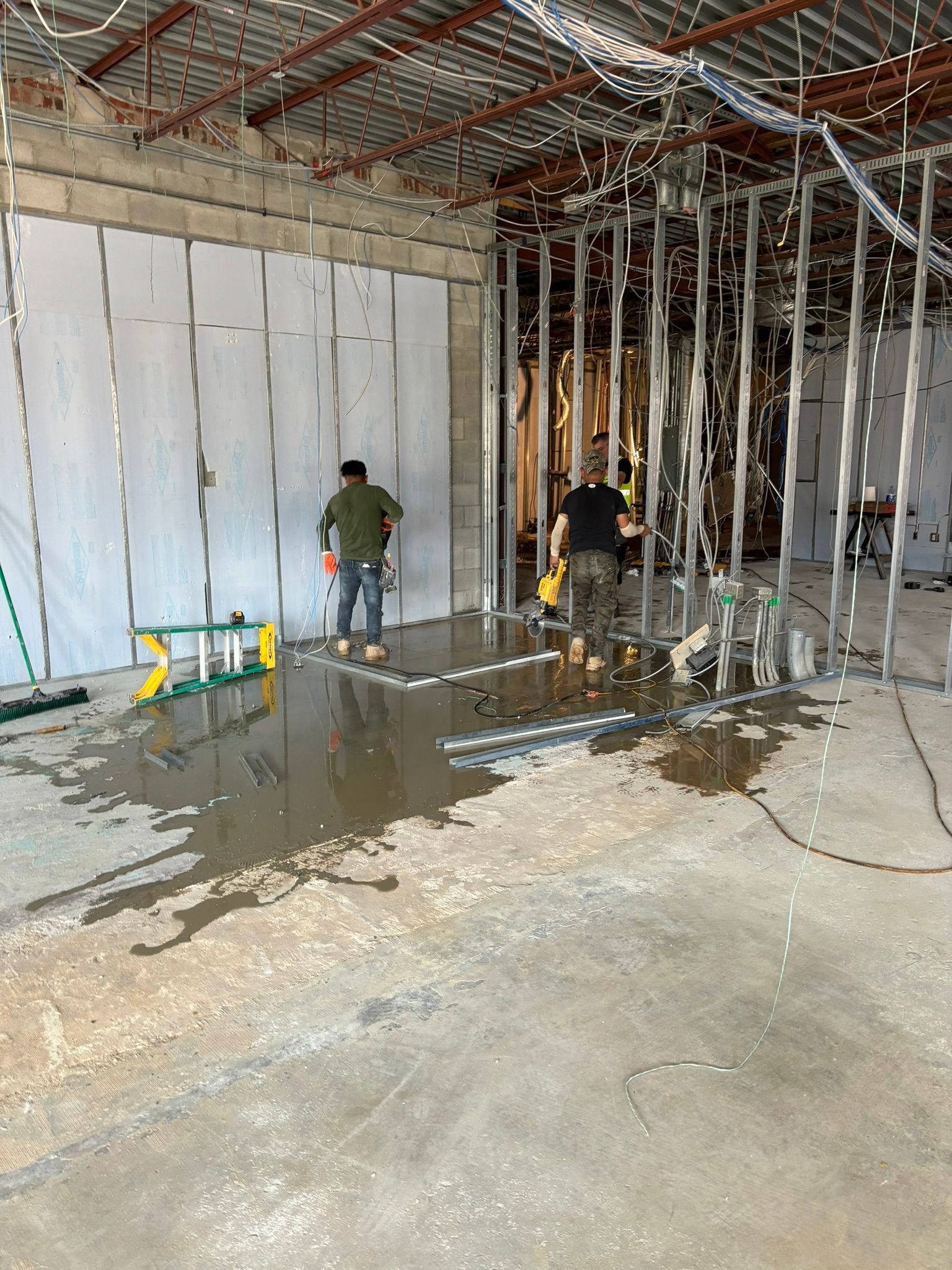 Construction site with two workers inside a building. One stands, the other crouches. Cement floor covered with water.