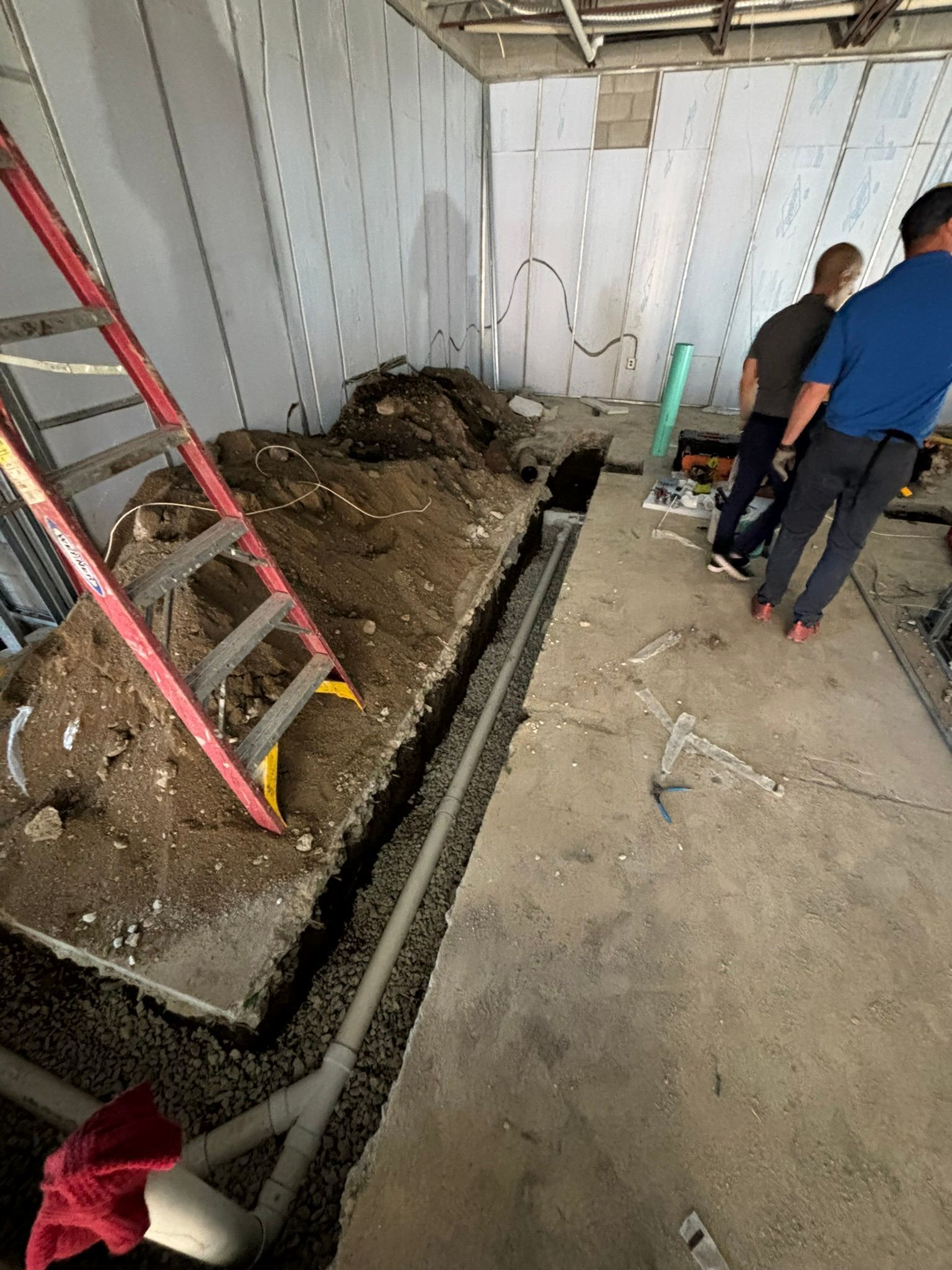 Construction site: Trench dug, men in blue and gray shirts, ladder, gray walls, concrete floor.