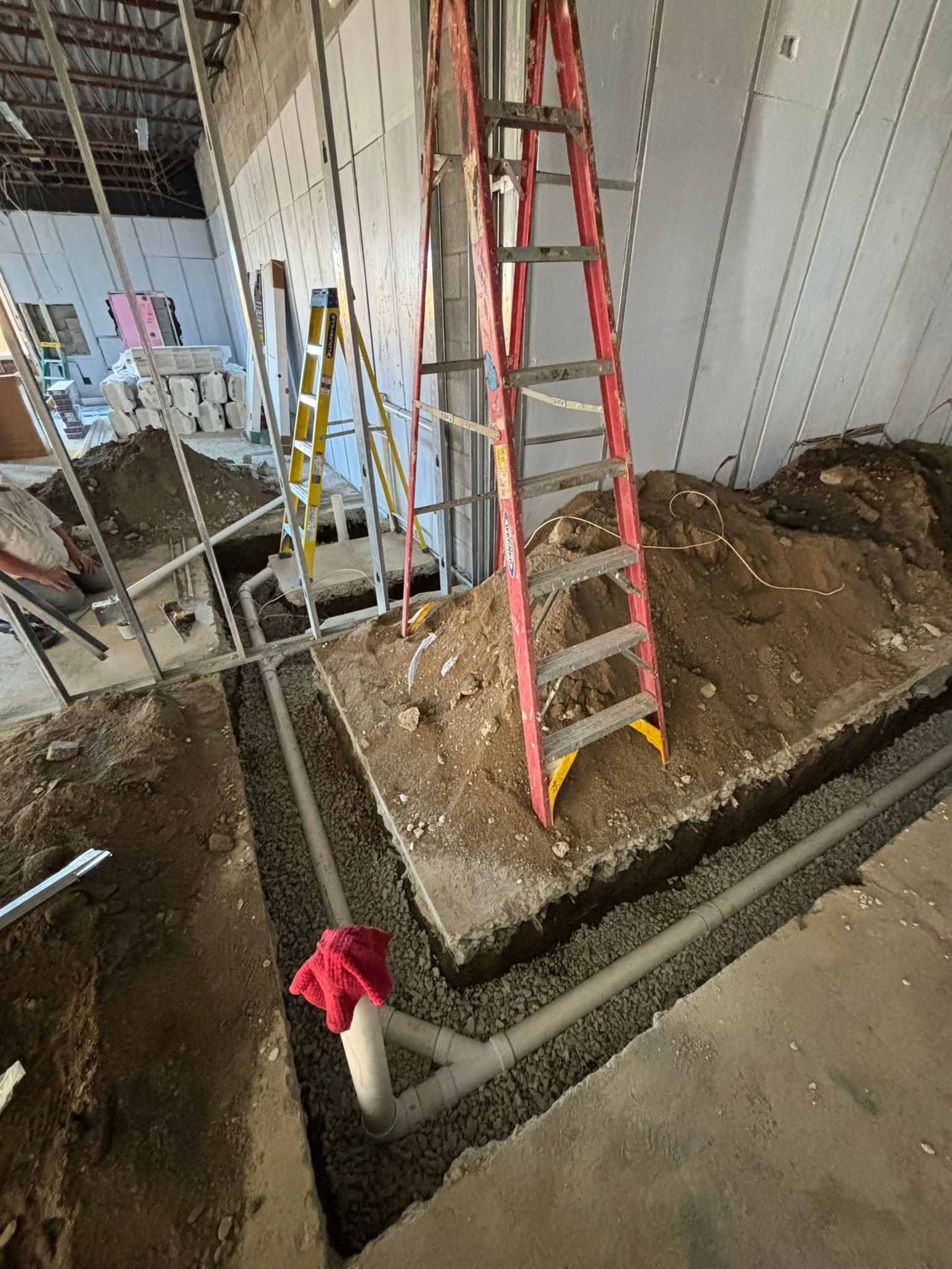 Construction site: red ladder in trench with gray piping, dirt, gravel, and white interior walls.