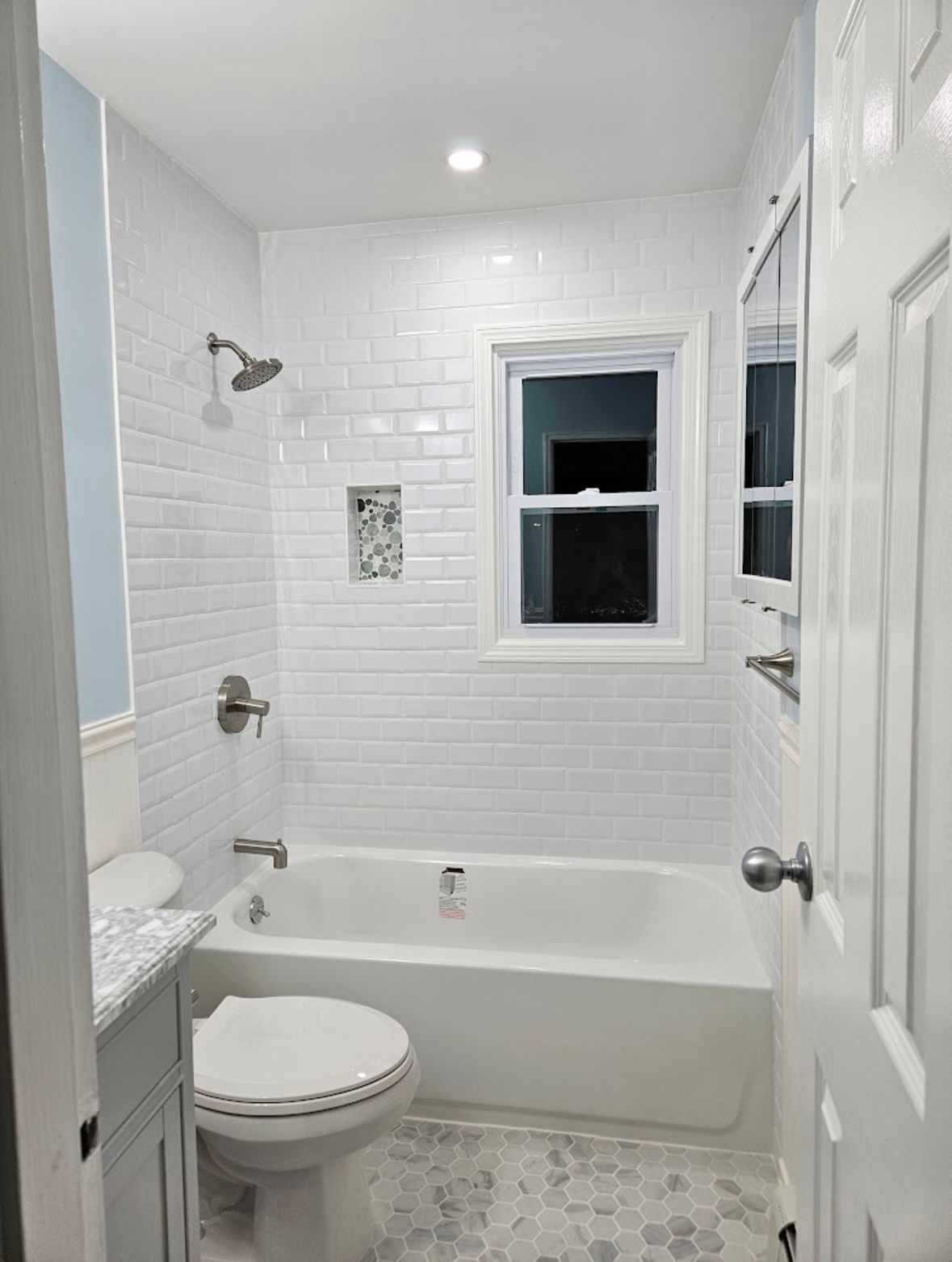 Small bathroom with white subway tile, a bathtub, and a toilet. A window provides natural light.
