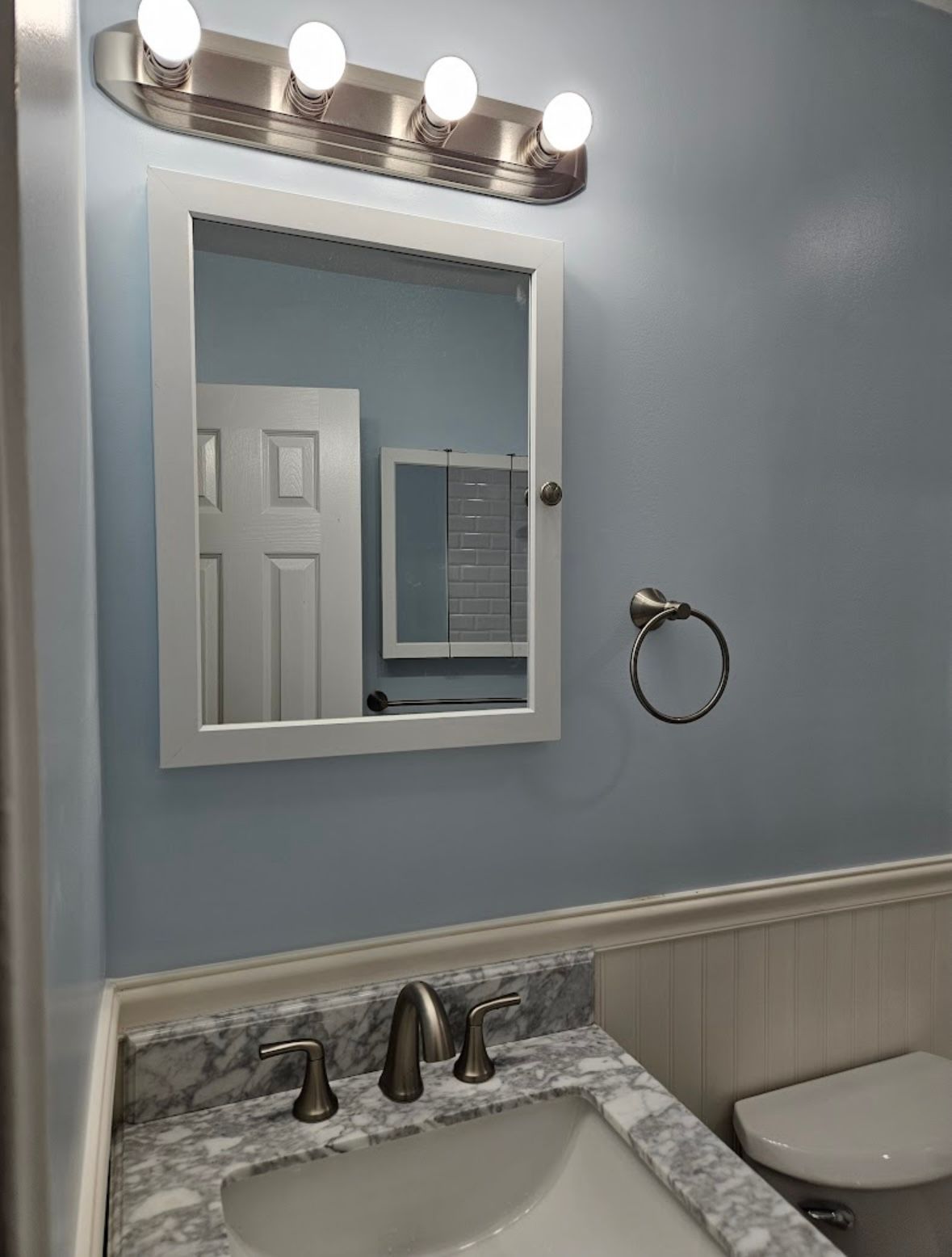 Bathroom with blue walls, a white-framed mirror, a vanity with a marble countertop, and a toilet.