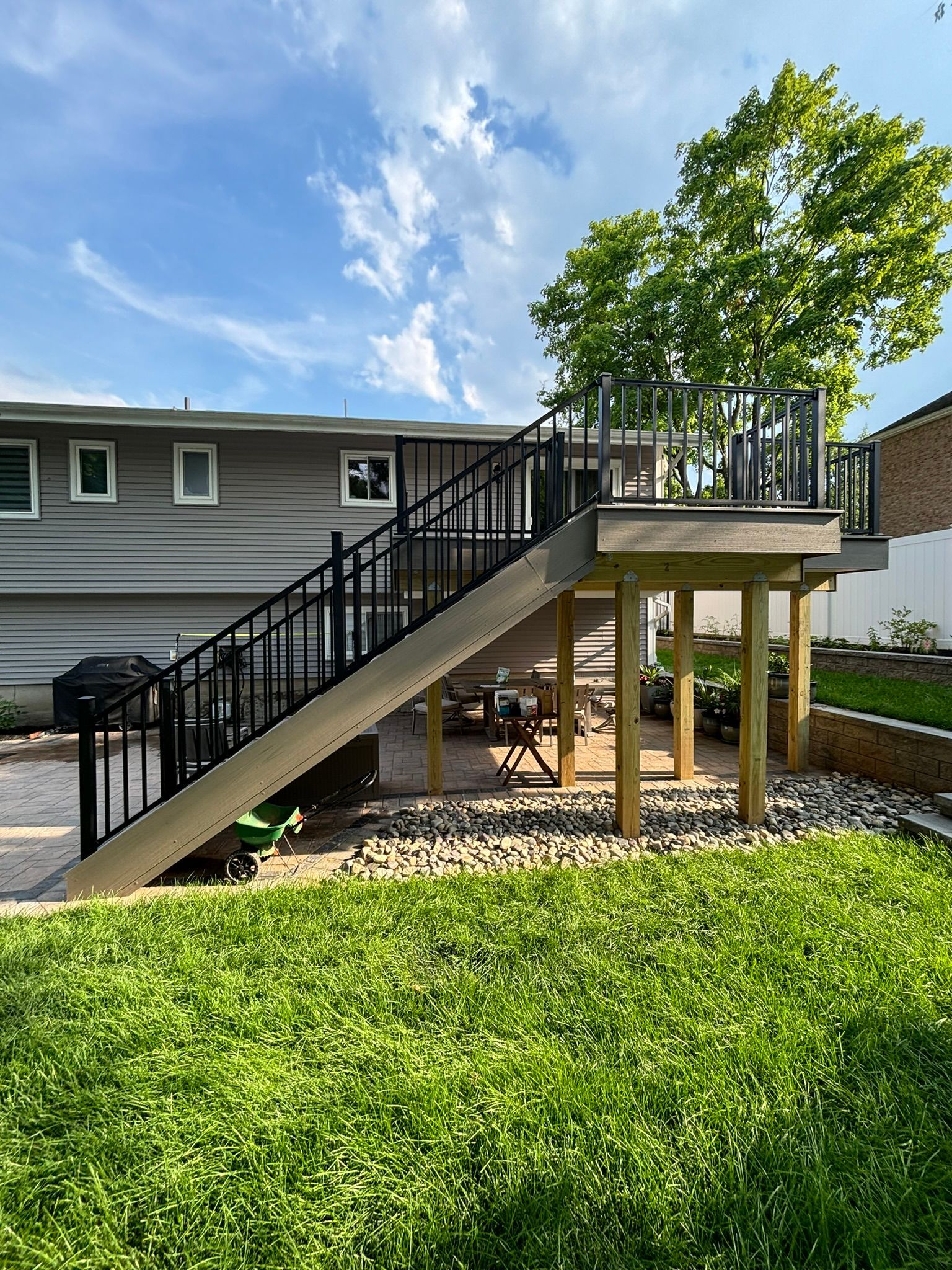 Deck with black railing and stairs, supported by wooden posts, with a green lawn and blue sky.