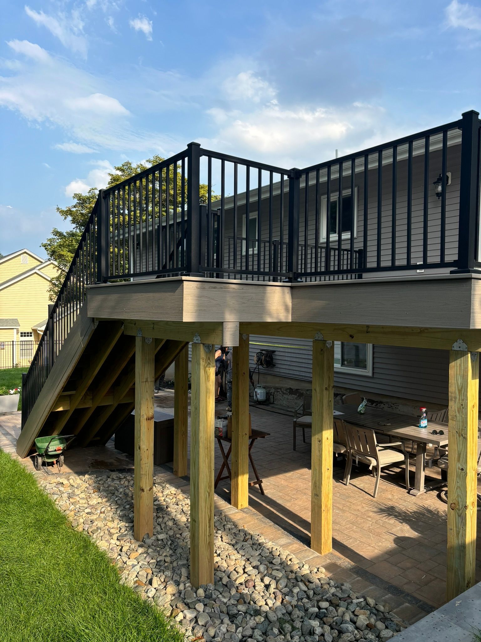 Elevated wooden deck with black railing, stairs, and support posts. Outdoor patio underneath; stone ground, grass.