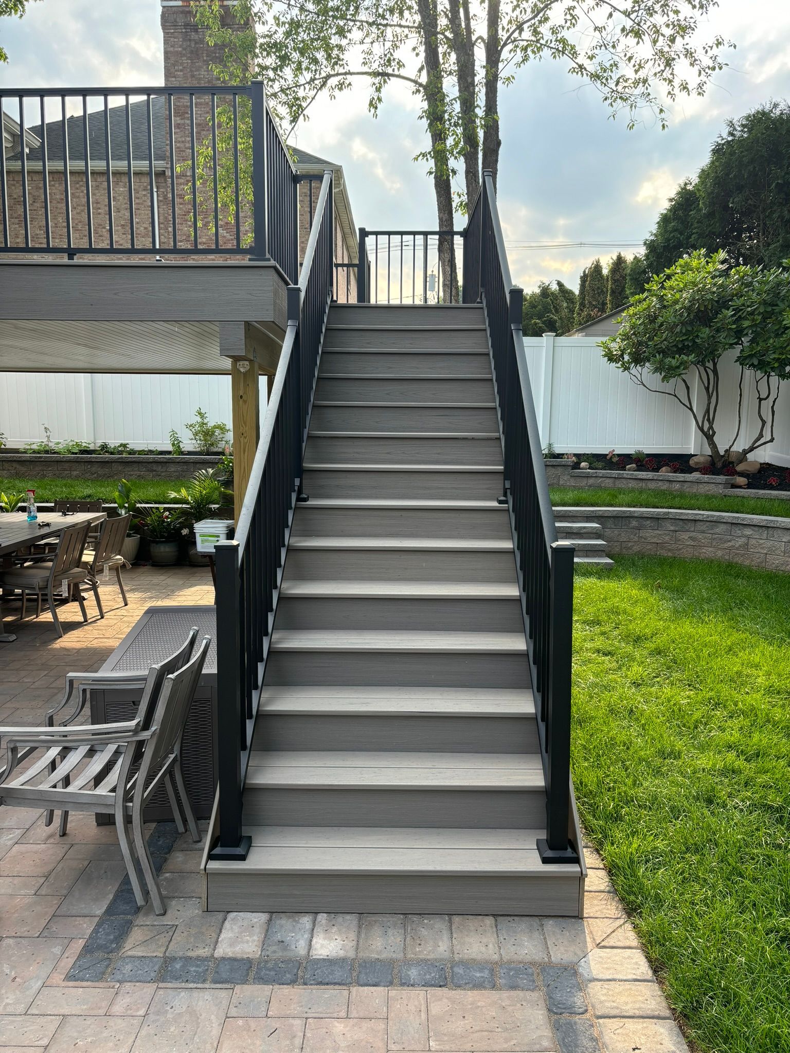 Gray composite stairs with black railing leading up to a deck. Backyard setting.