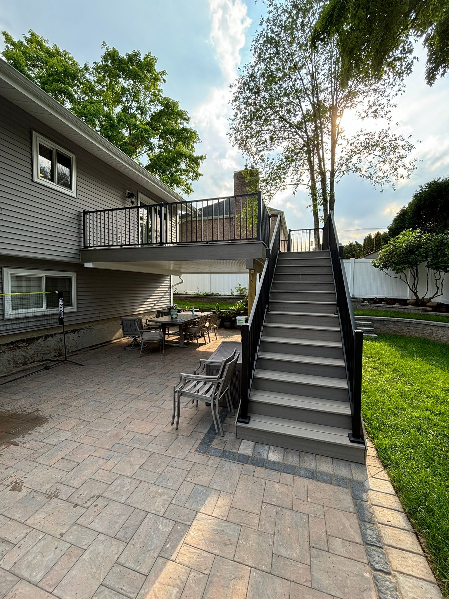 Backyard with a deck, stairs, and a patio.  Gray house, black railing, and brick patio with outdoor furniture.