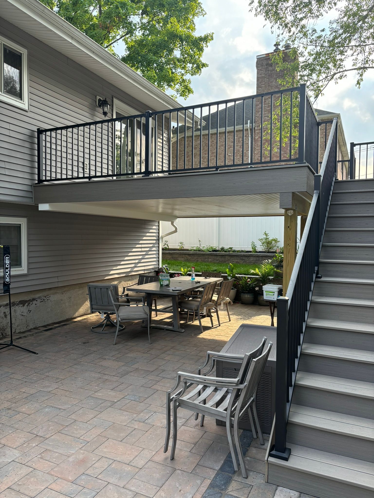 Backyard patio with dining set, beneath a raised deck with stairs and black railing.