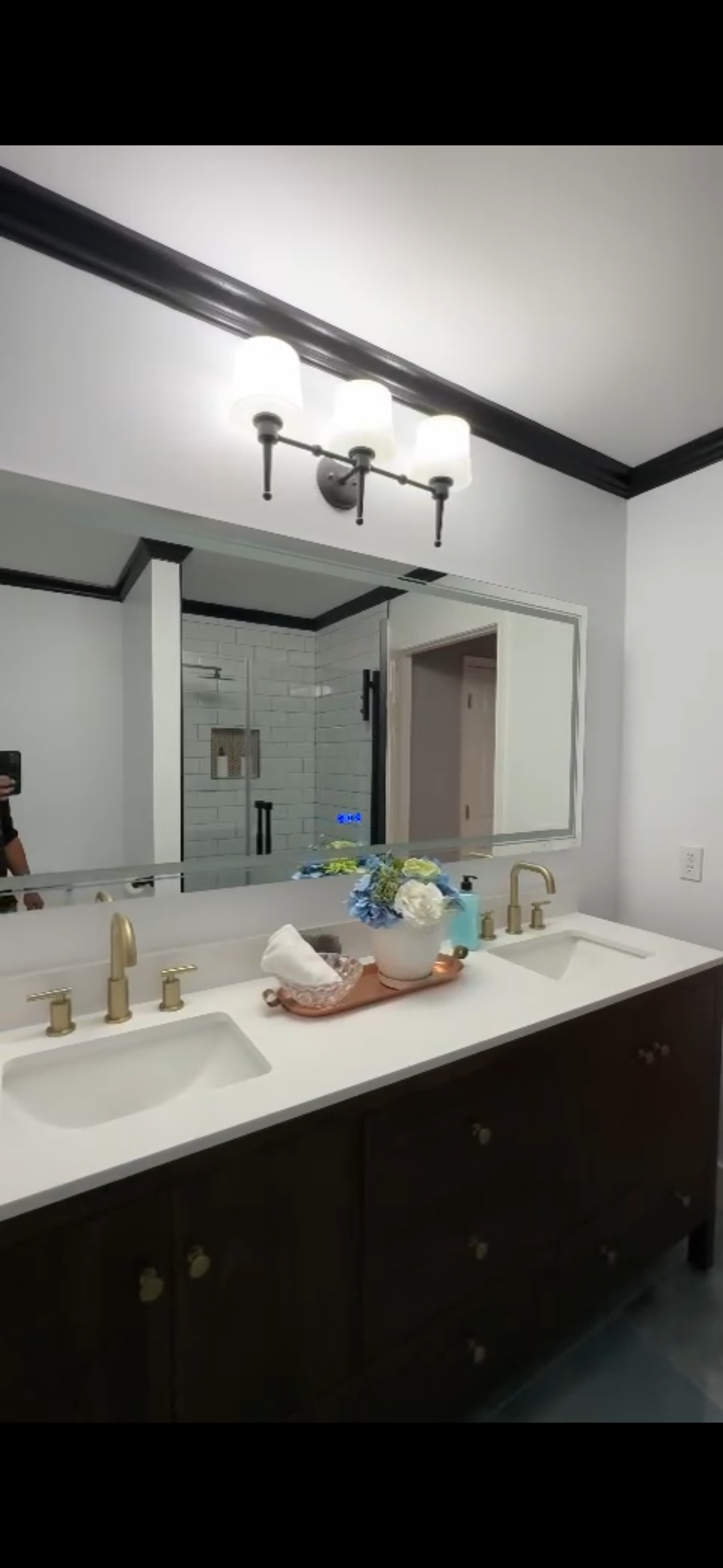 Bathroom with double sinks, large mirror, overhead lighting, and dark cabinetry. White walls with black trim.