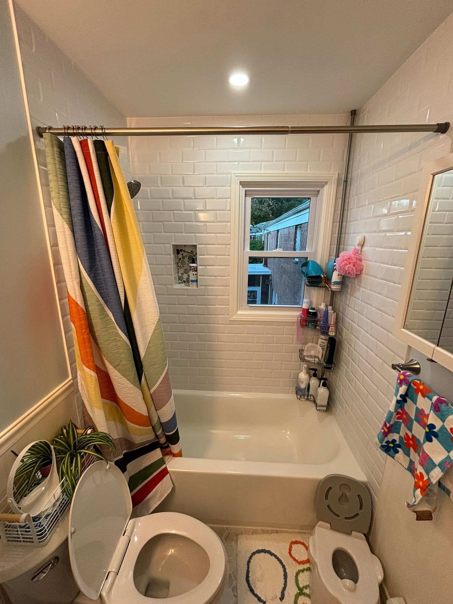 Small bathroom with a tub, toilet, window, colorful shower curtain, and various toiletries.