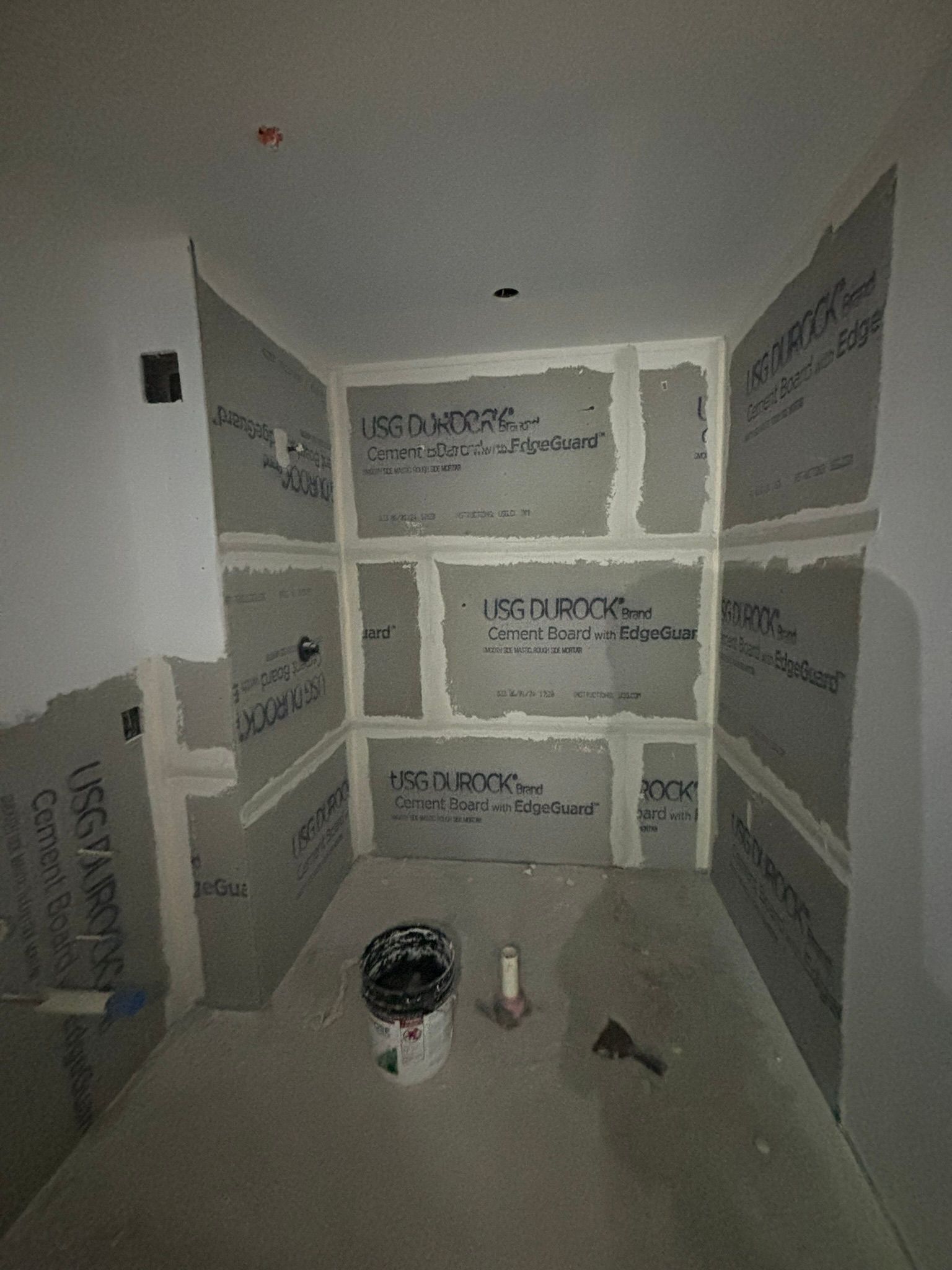 Shower stall under construction, gray cement boards installed on walls, bucket and tools on floor.