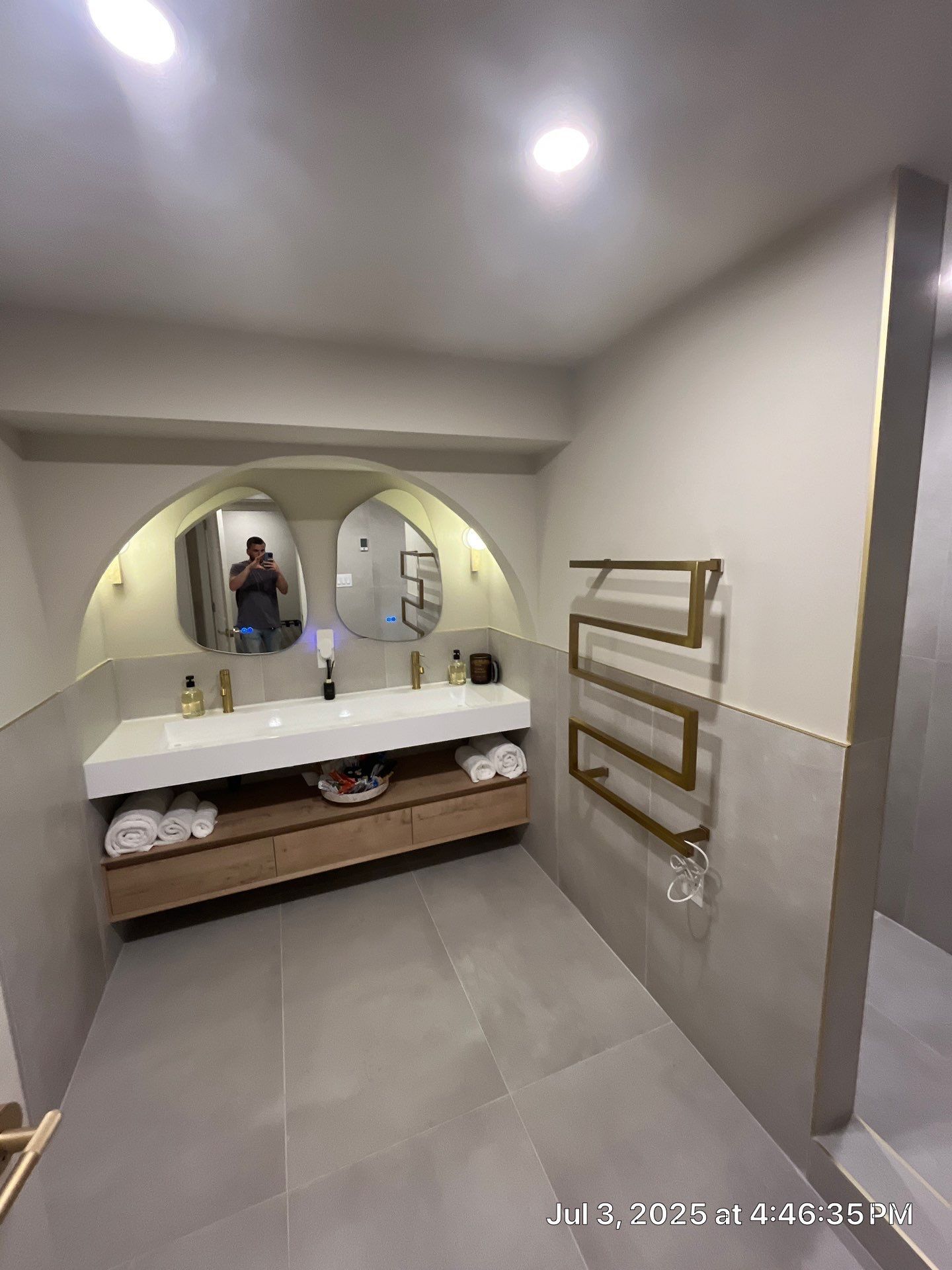 Modern bathroom with floating double vanity, oval mirrors, gold towel rack, and gray tile floor.